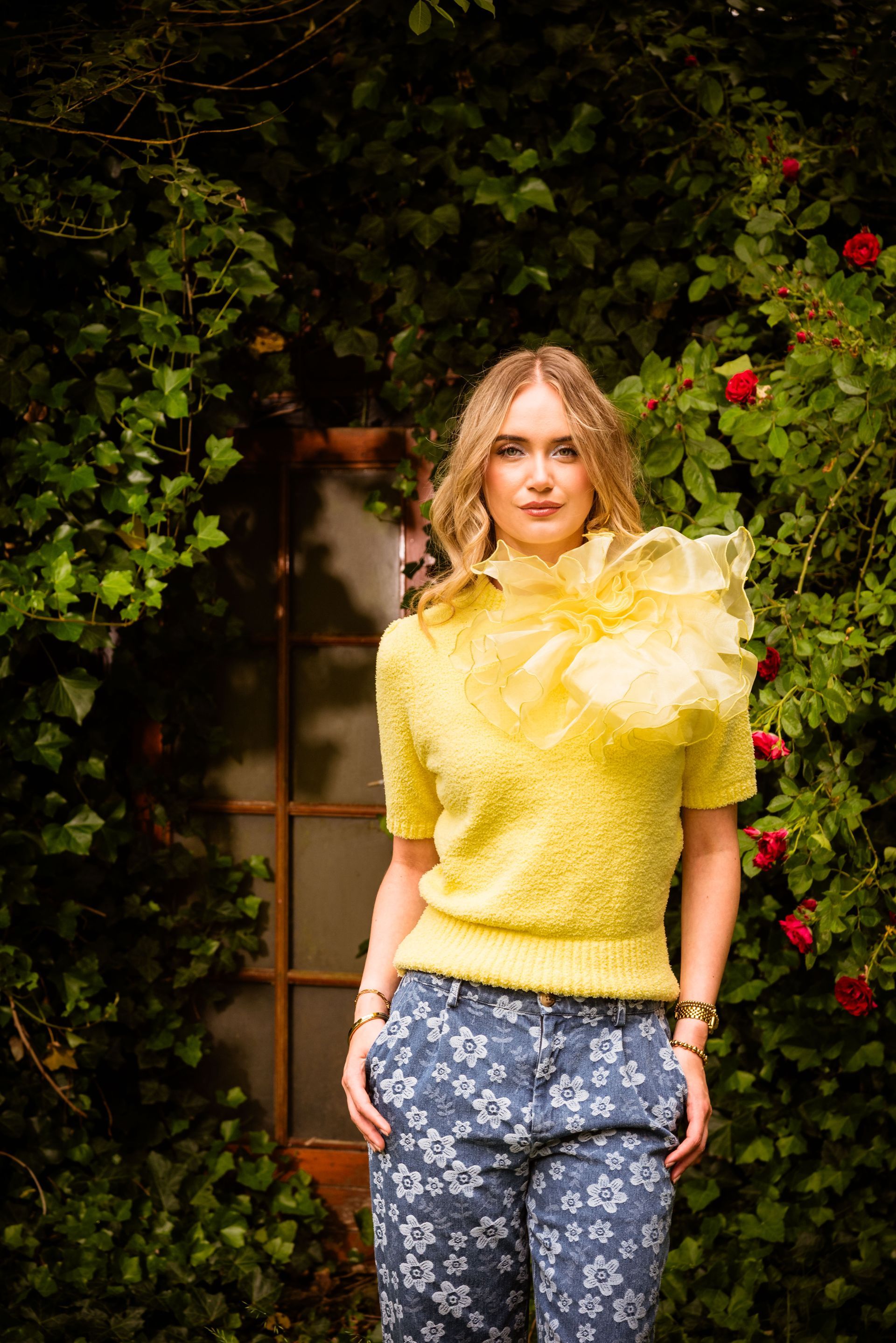 Een vrouw in een gele top en een broek met bloemenprint poseert buiten bij een raam dat is omgeven door groen en rozen.