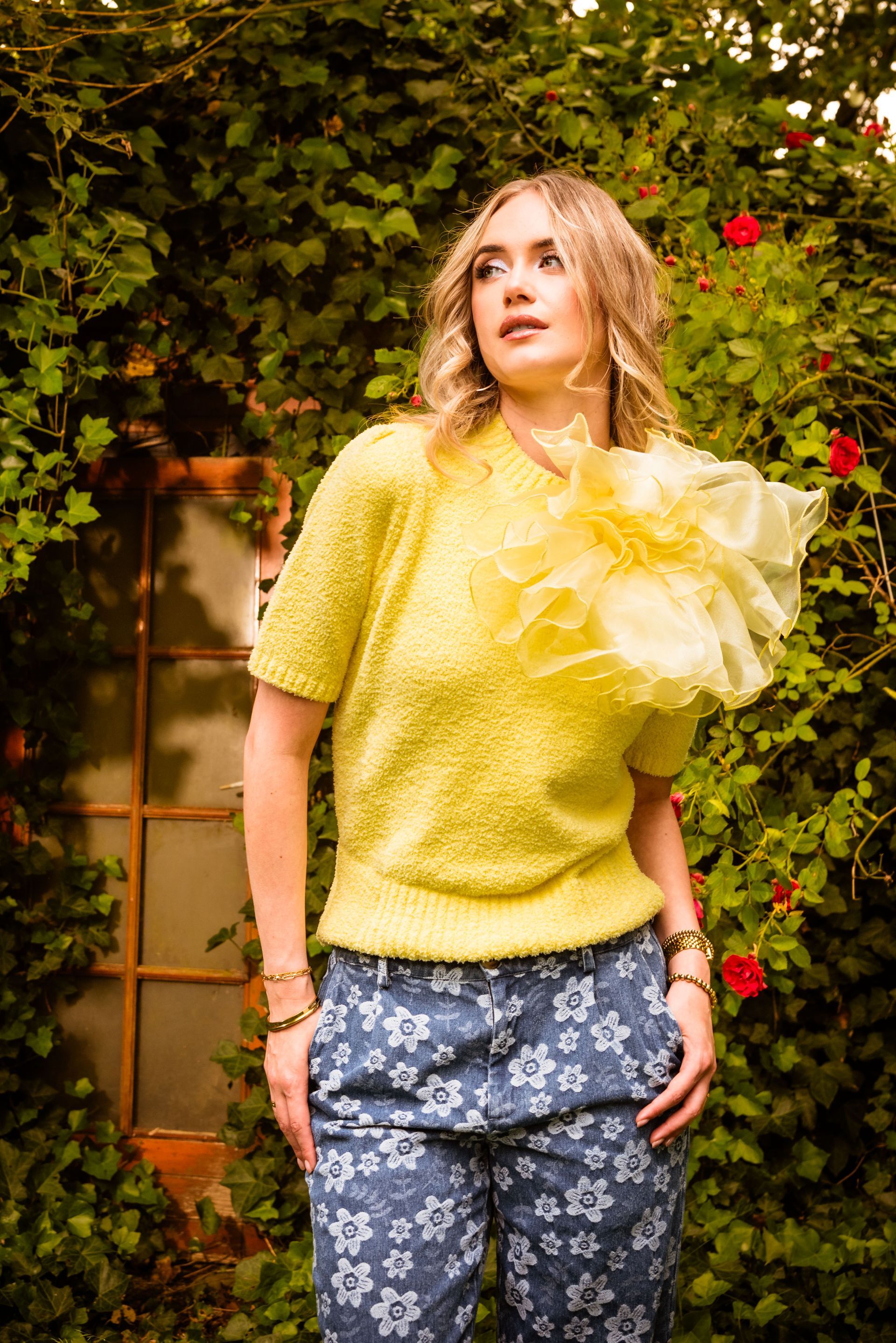 Een vrouw in een gele trui en een blauwe broek met bloemenprint staat voor een met groene klimop begroeide muur.