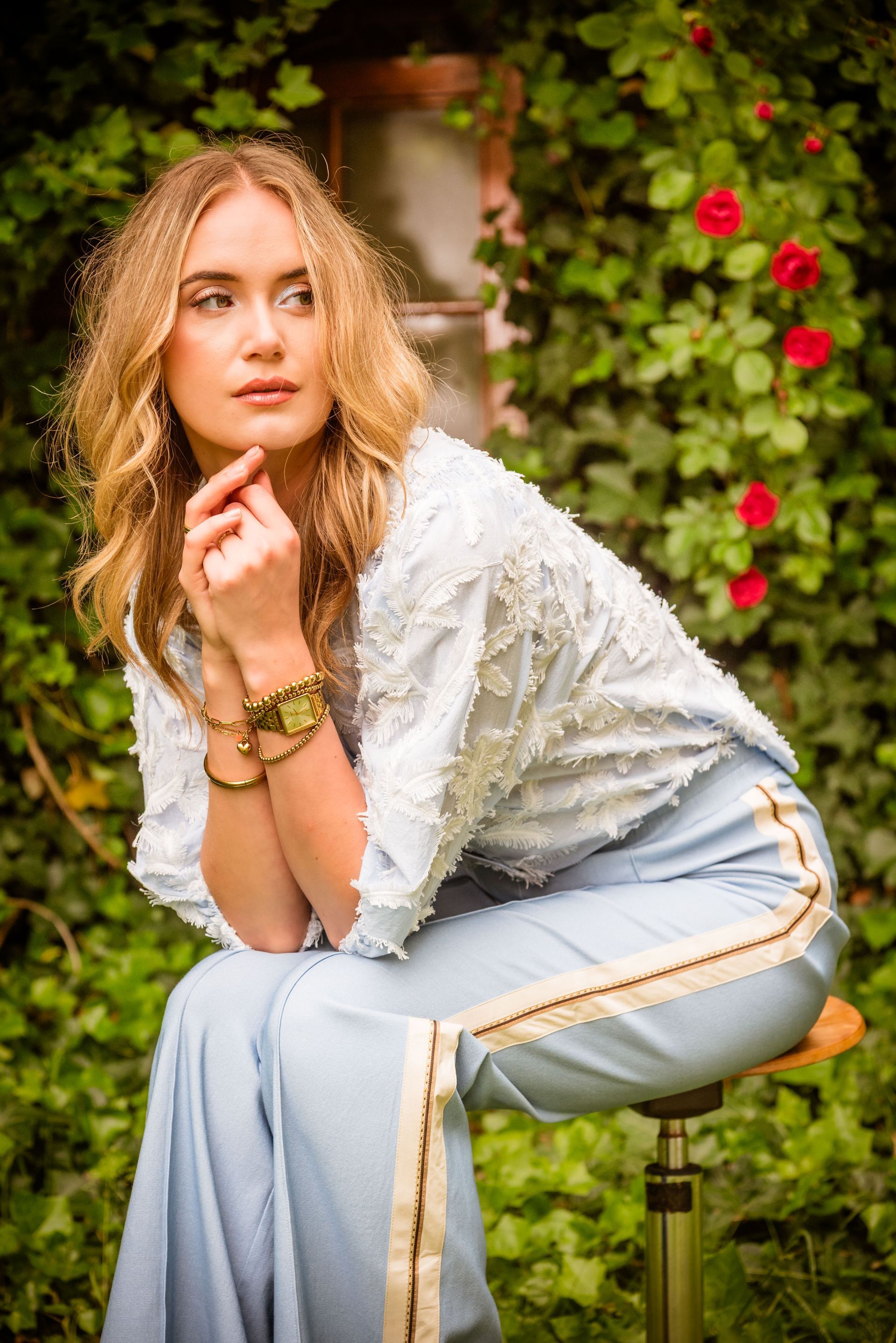 Vrouw in blauwe outfit zit, haar kin rustend op haar hand, en kijkt niet in beeld. Klimop en rozen op de achtergrond.