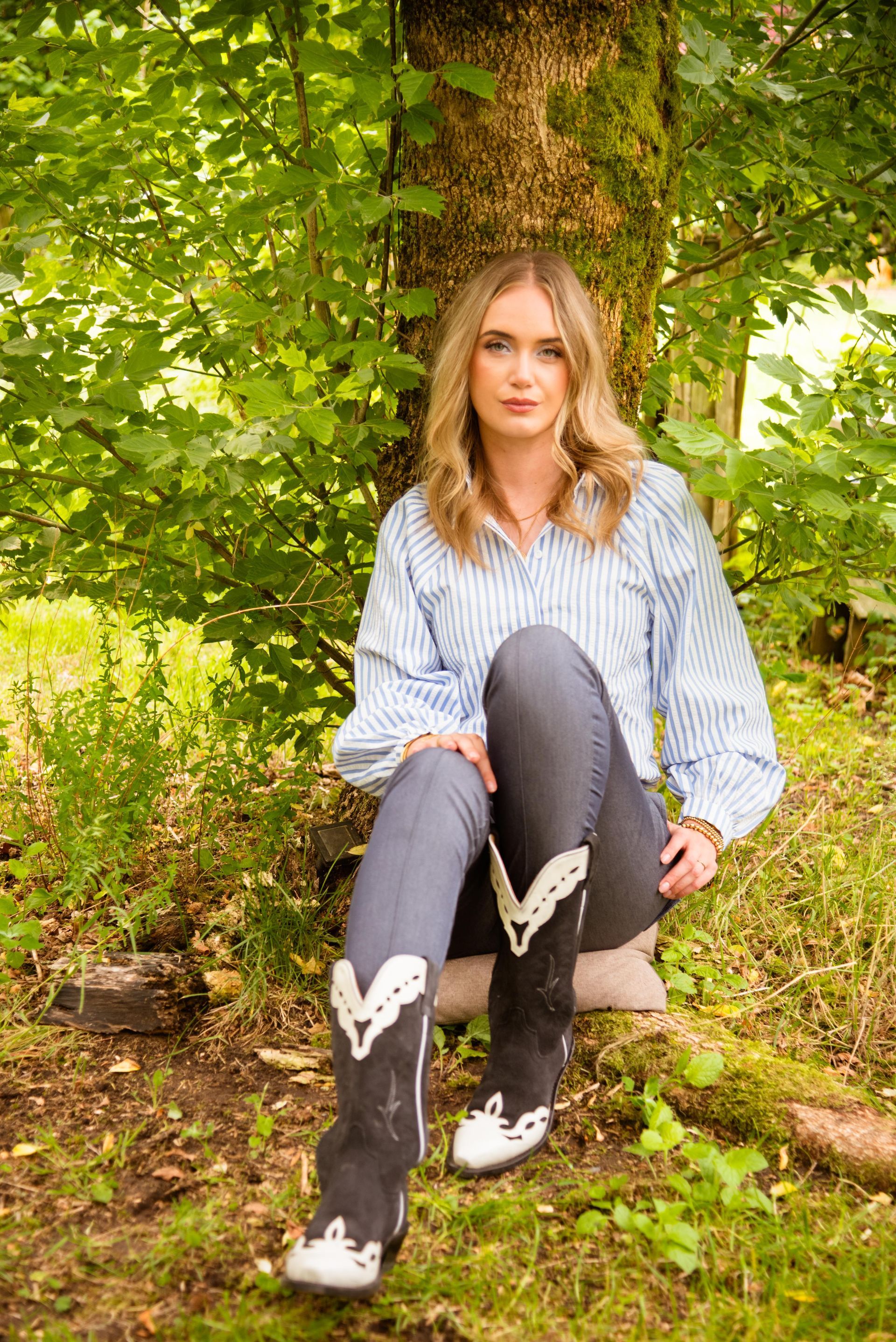 Een vrouw in een blauw gestreept shirt en jeans zit bij een boom, draagt cowboylaarzen en kijkt voor zich uit.