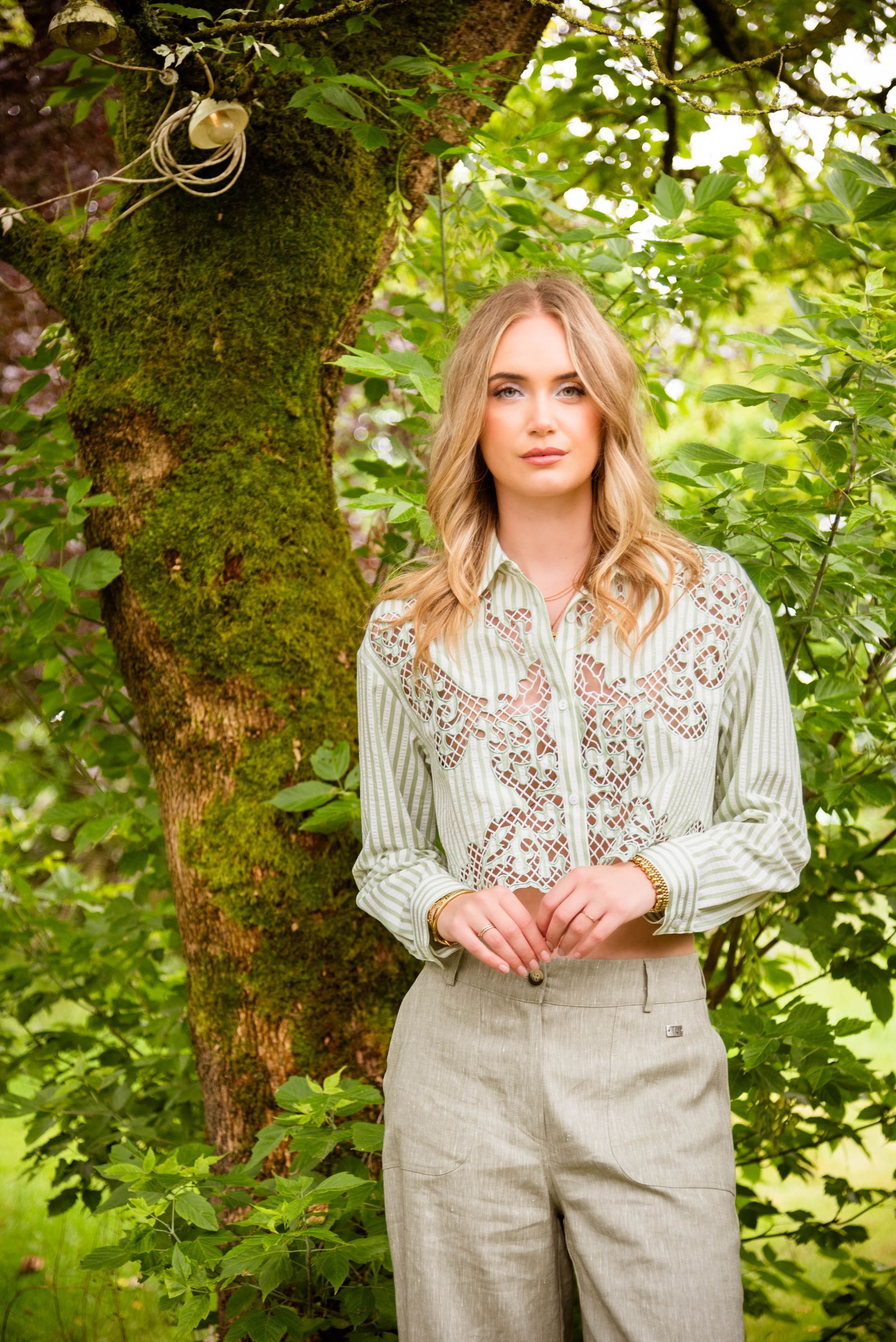Vrouw in geborduurd shirt en linnen broek, staand naast een met mos begroeide boom in een weelderig groene omgeving.