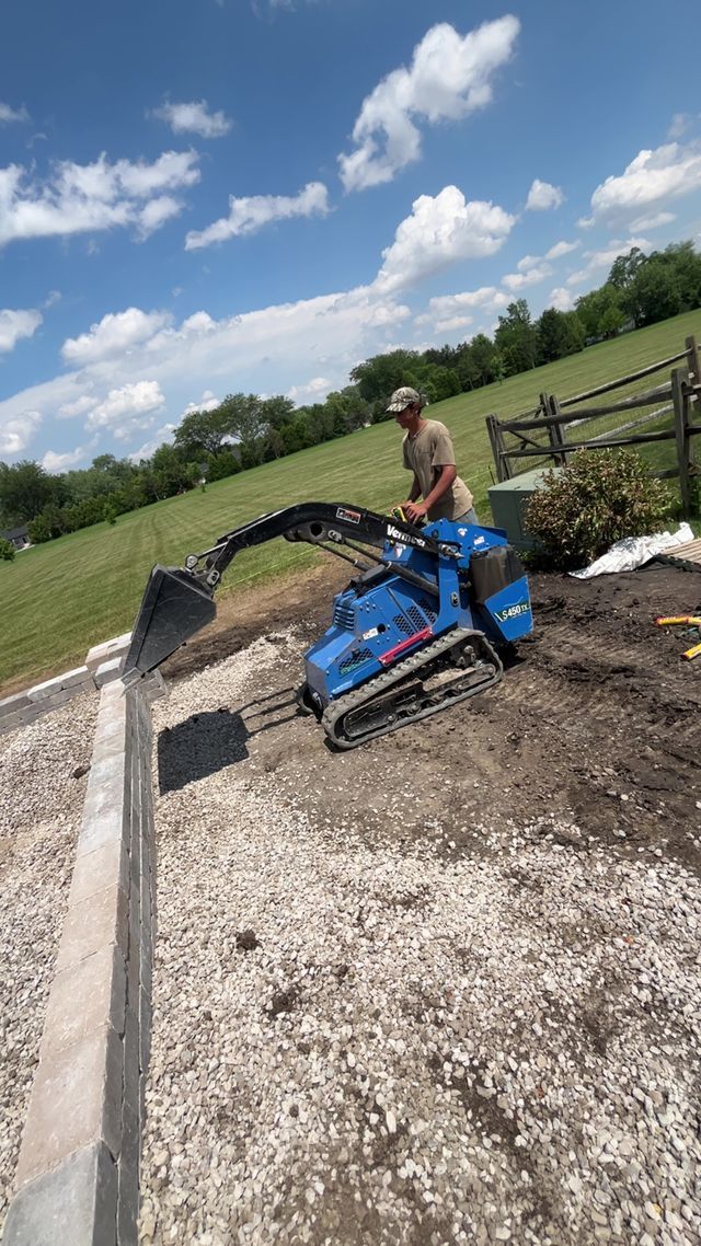 Man operating blue mini excavator on gravel path near a fence and grassy hill on a sunny day.