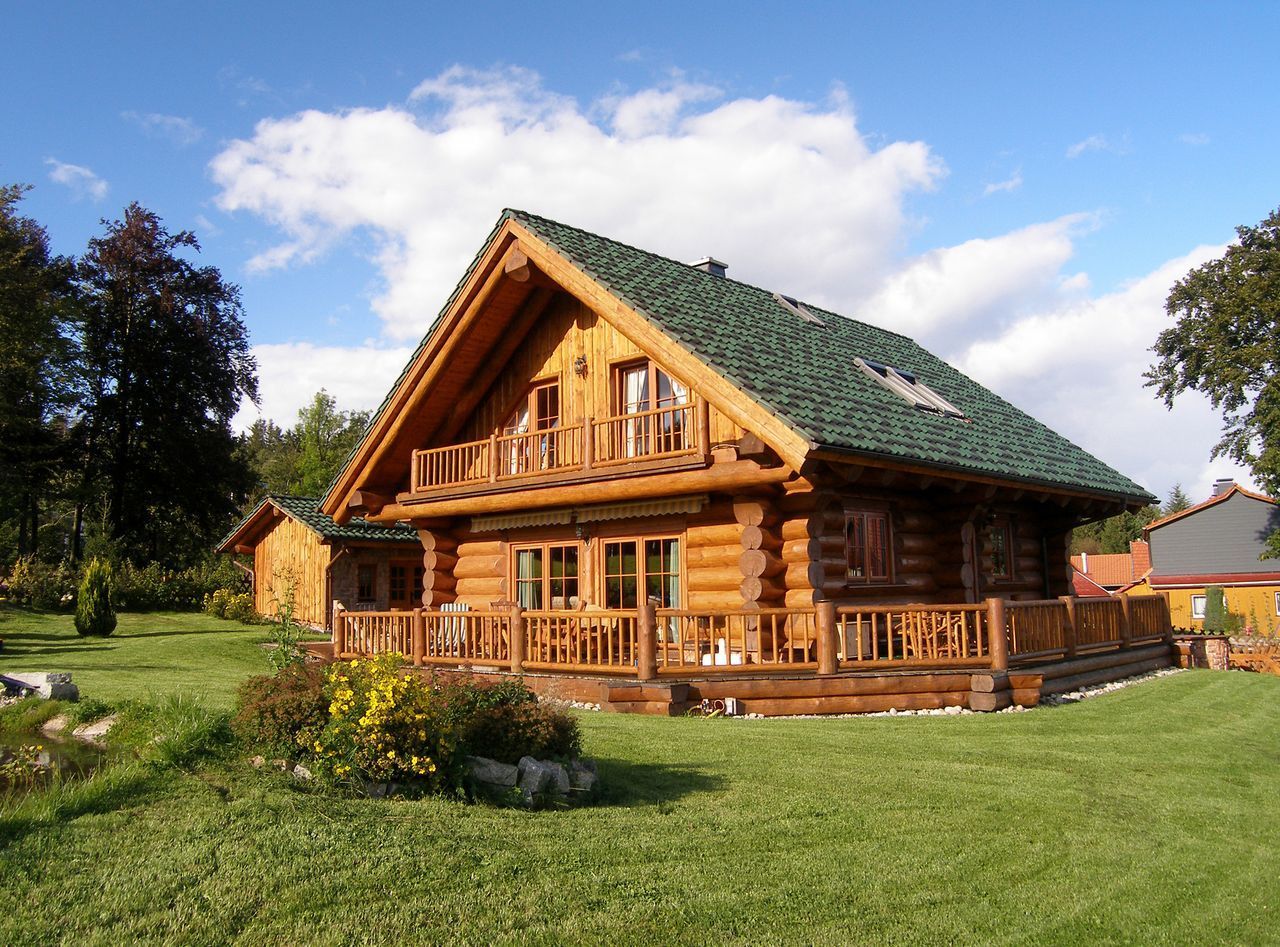 A two-story log cabin with a green roof and a wrap-around wooden porch, set in a sunny, grassy yard with trees nearby.