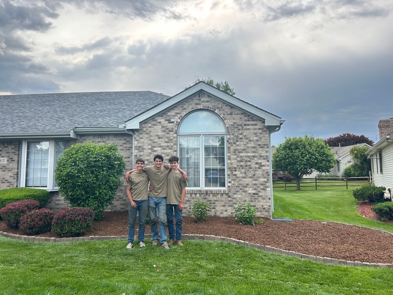 Three people standing in front of a house, arms around each other. Overcast sky.