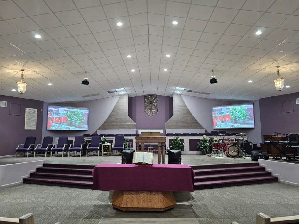 A view of a church sanctuary with purple walls, rows of chairs on a raised stage, two large screens, and a central altar.