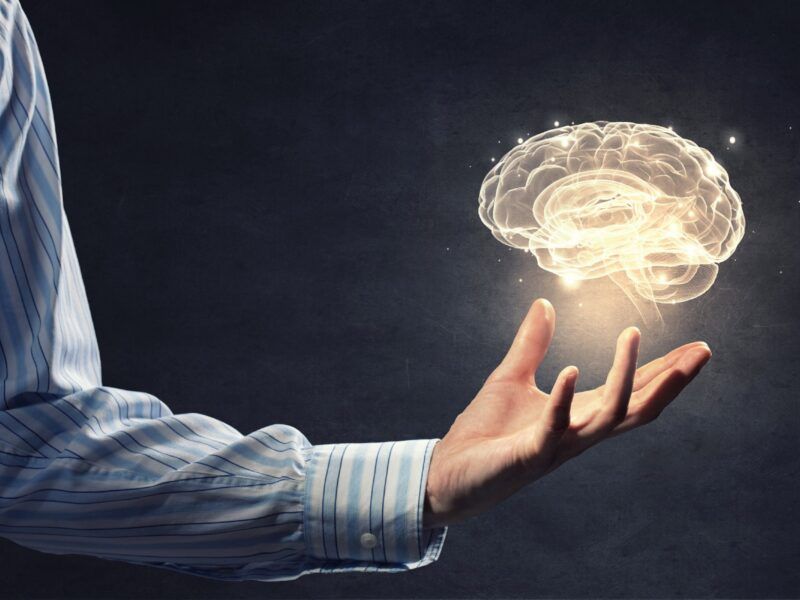 A hand in a blue striped shirt holds a glowing, translucent 3D human brain against a dark, textured background.