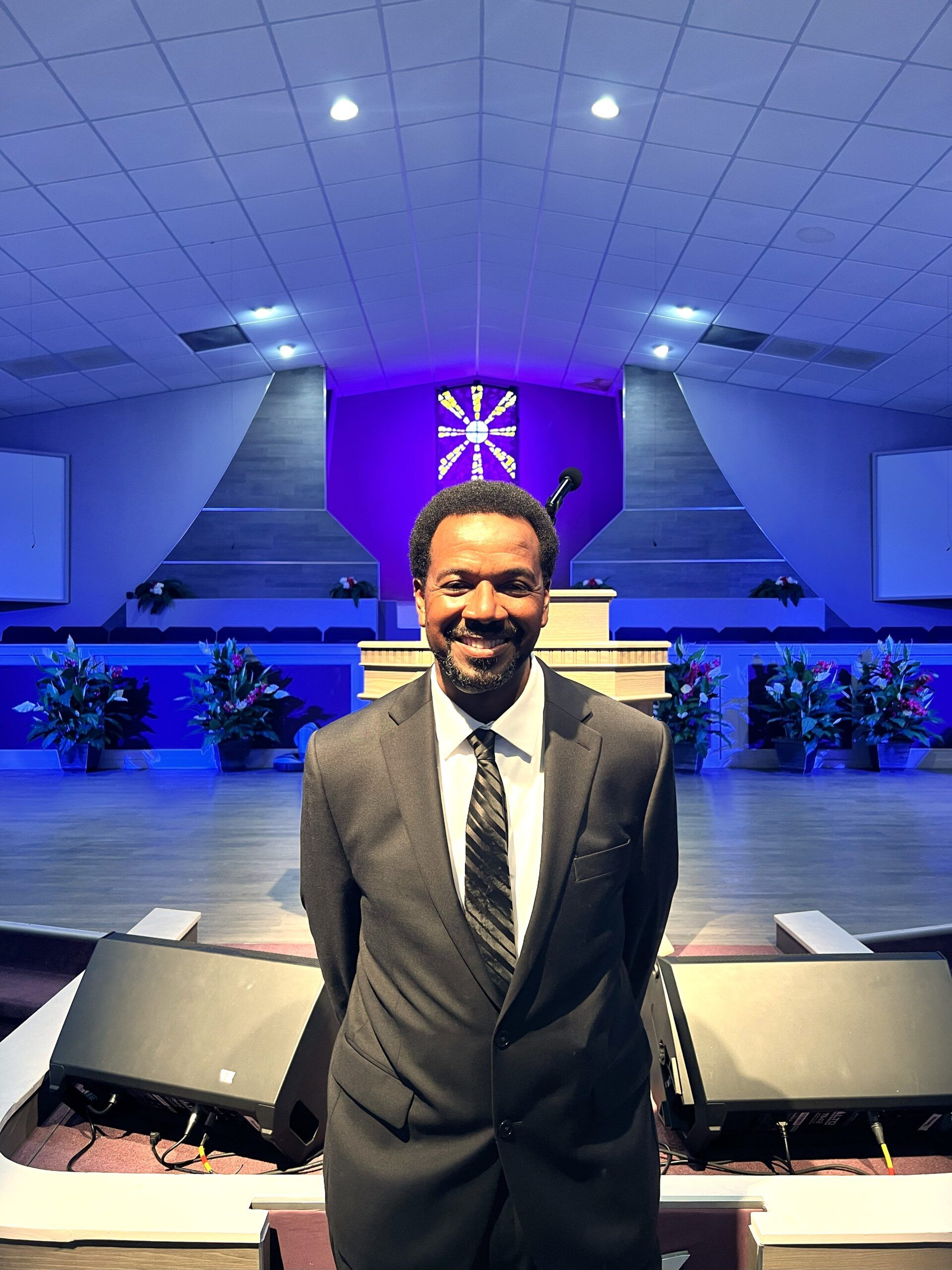 A smiling man in a suit and tie stands on a church stage with purple lighting, flowers, and a cross in the background.