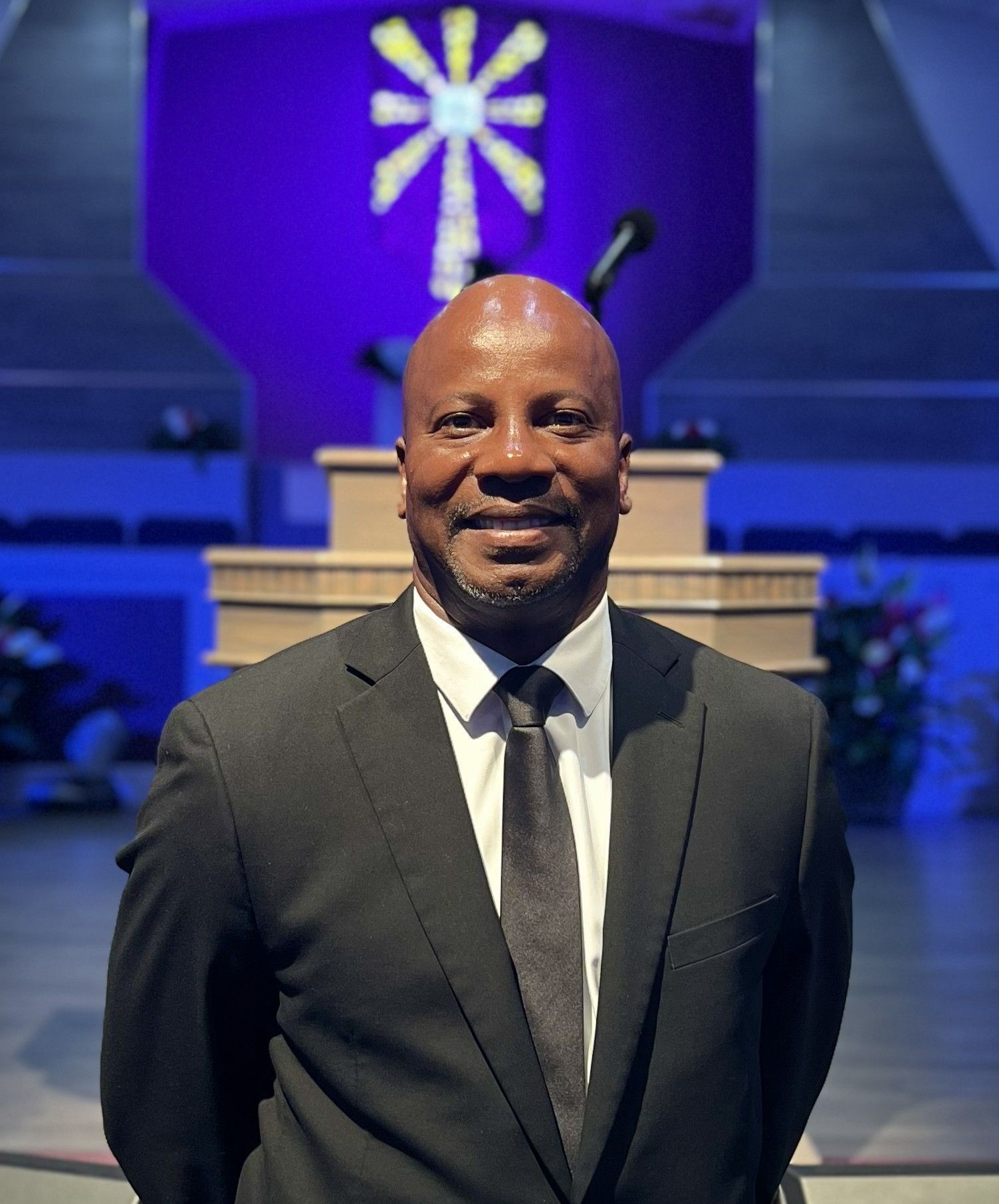 A person in a suit and tie stands in front of a church pulpit with a purple backdrop and a cross.