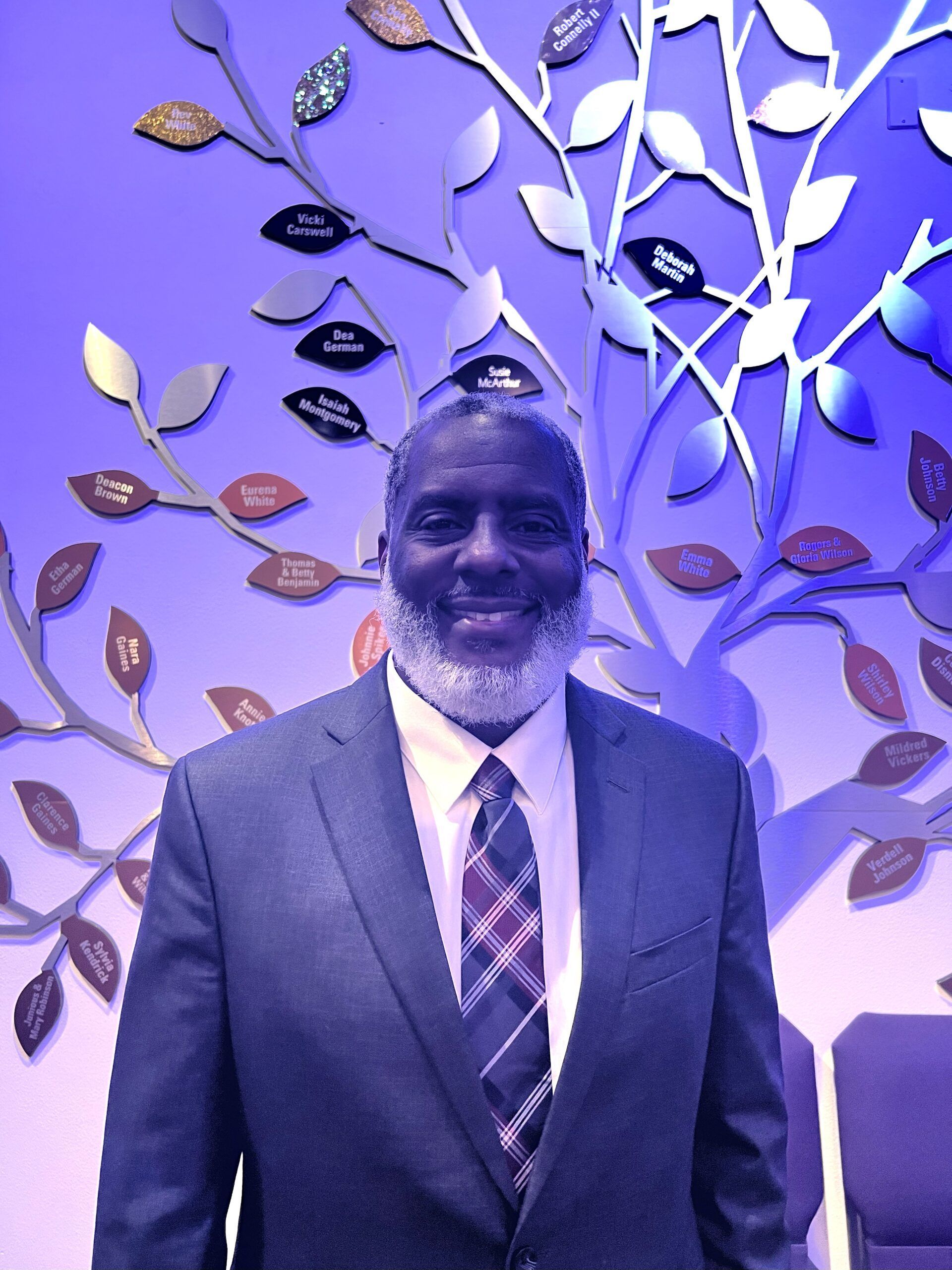 A smiling person wearing a suit and tie stands before a decorative metal tree wall sculpture with purple ambient lighting.