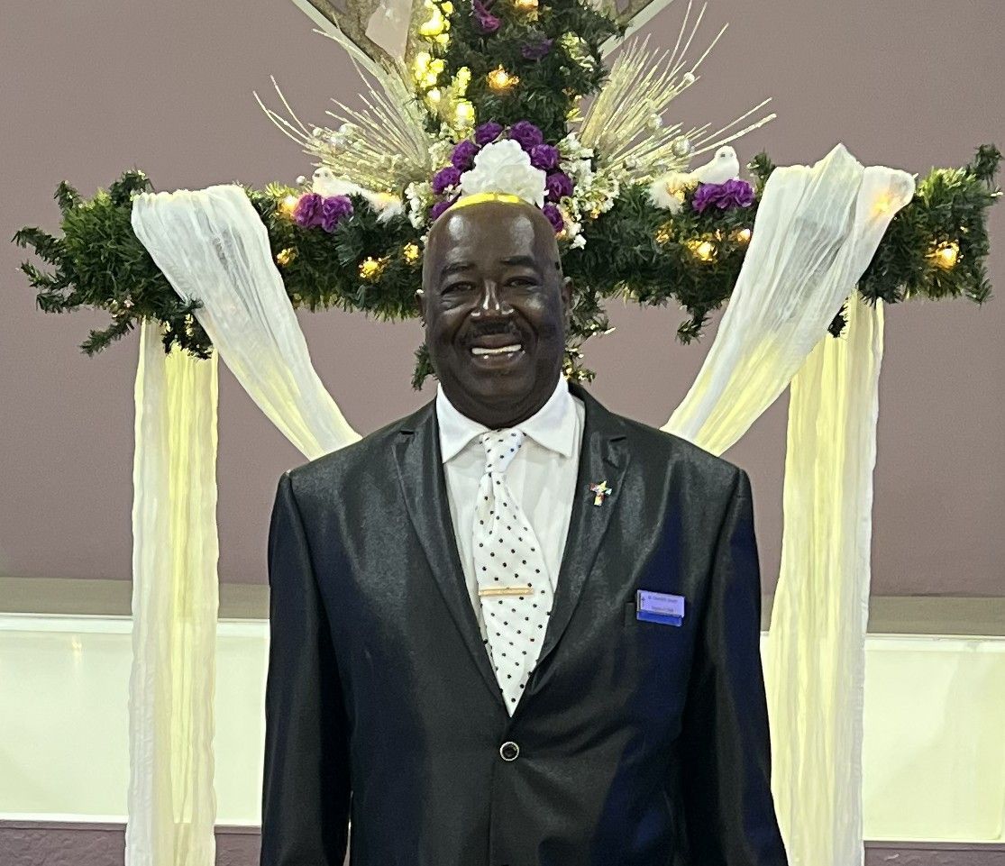 A smiling person in a dark suit and spotted tie standing in front of a decorated cross and flowing white fabric.