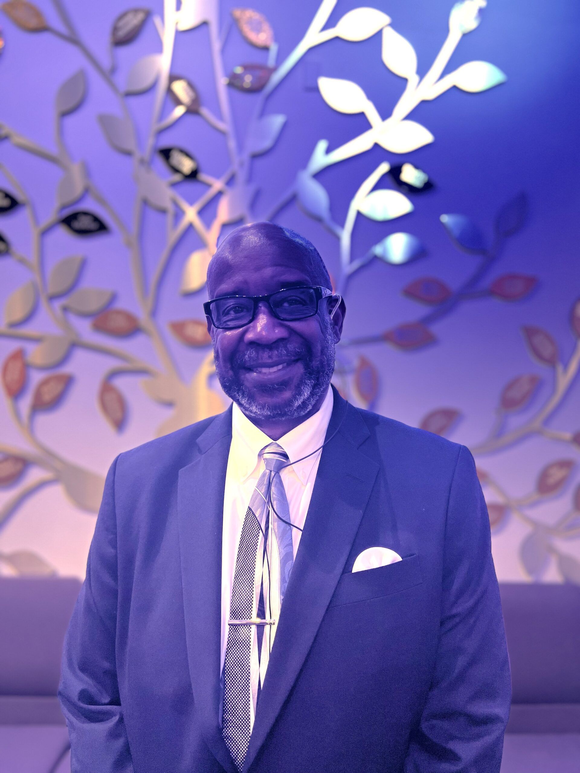 A person wearing a suit, white shirt, and patterned tie, smiling in front of a decorative, illuminated wall tree display.