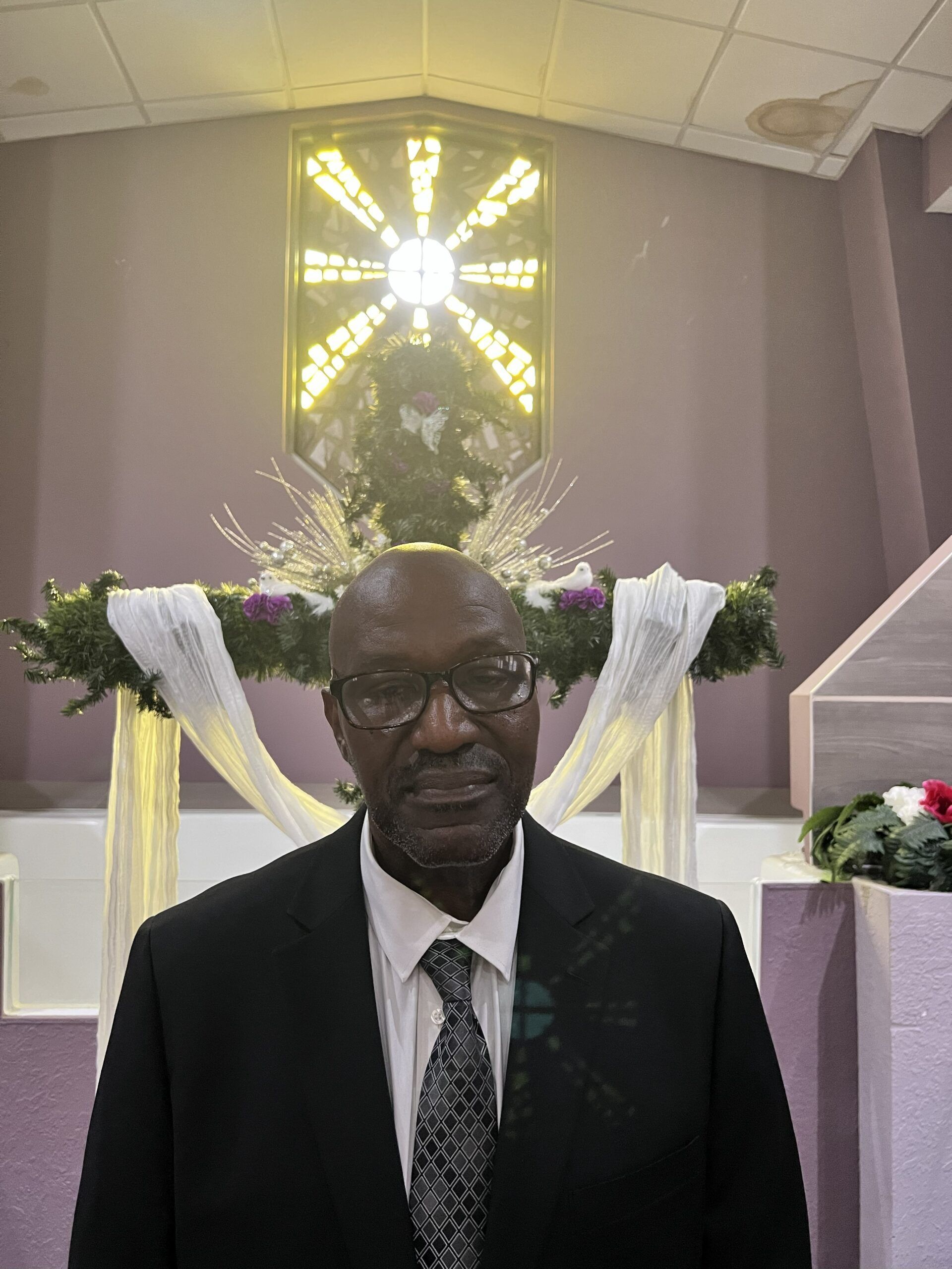 A person wearing a dark suit and patterned tie stands in front of a church altar decorated with flowers and white fabric.