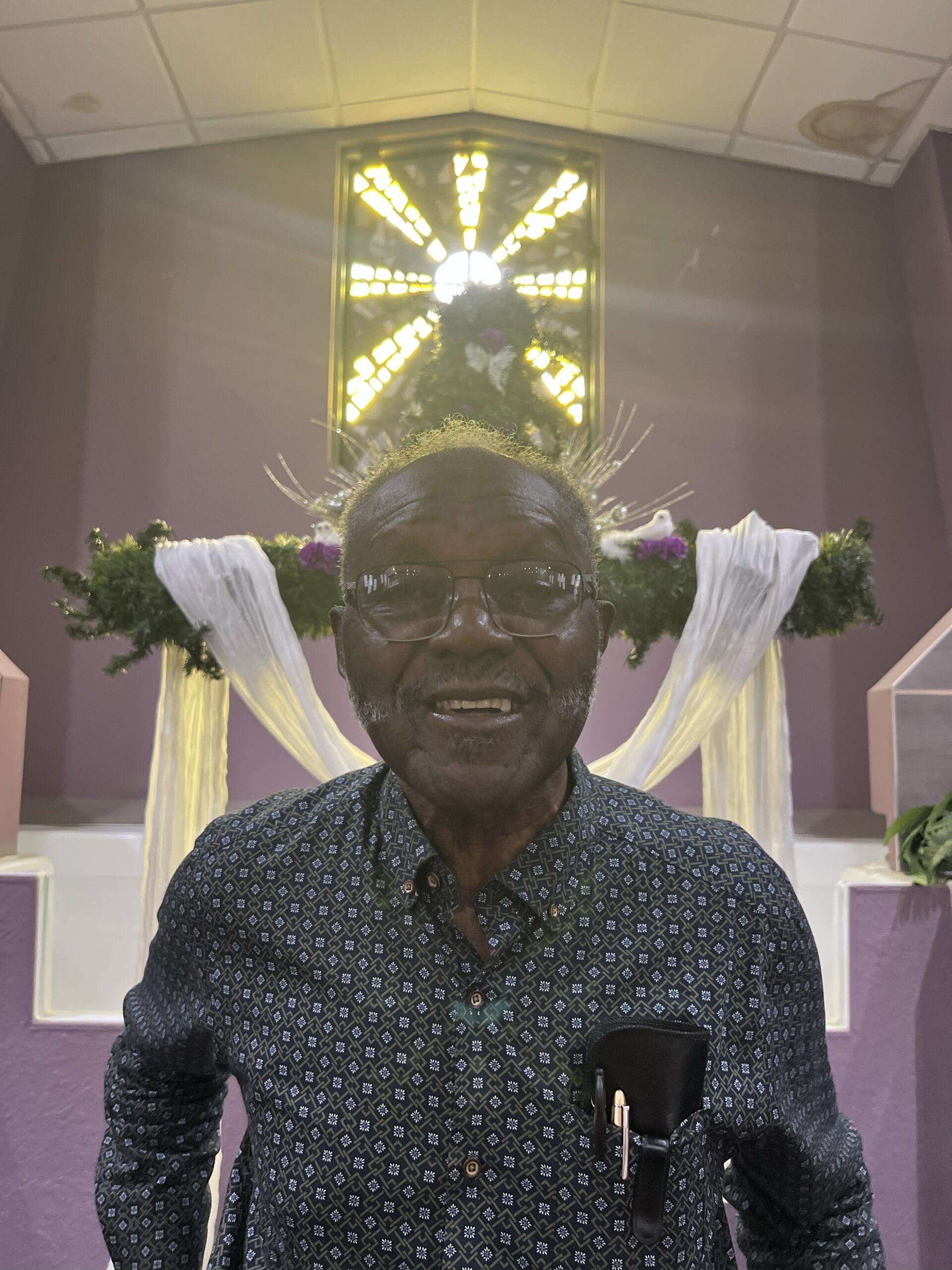 A smiling individual wearing a small crown stands in front of a church altar decorated with white fabric and greenery.