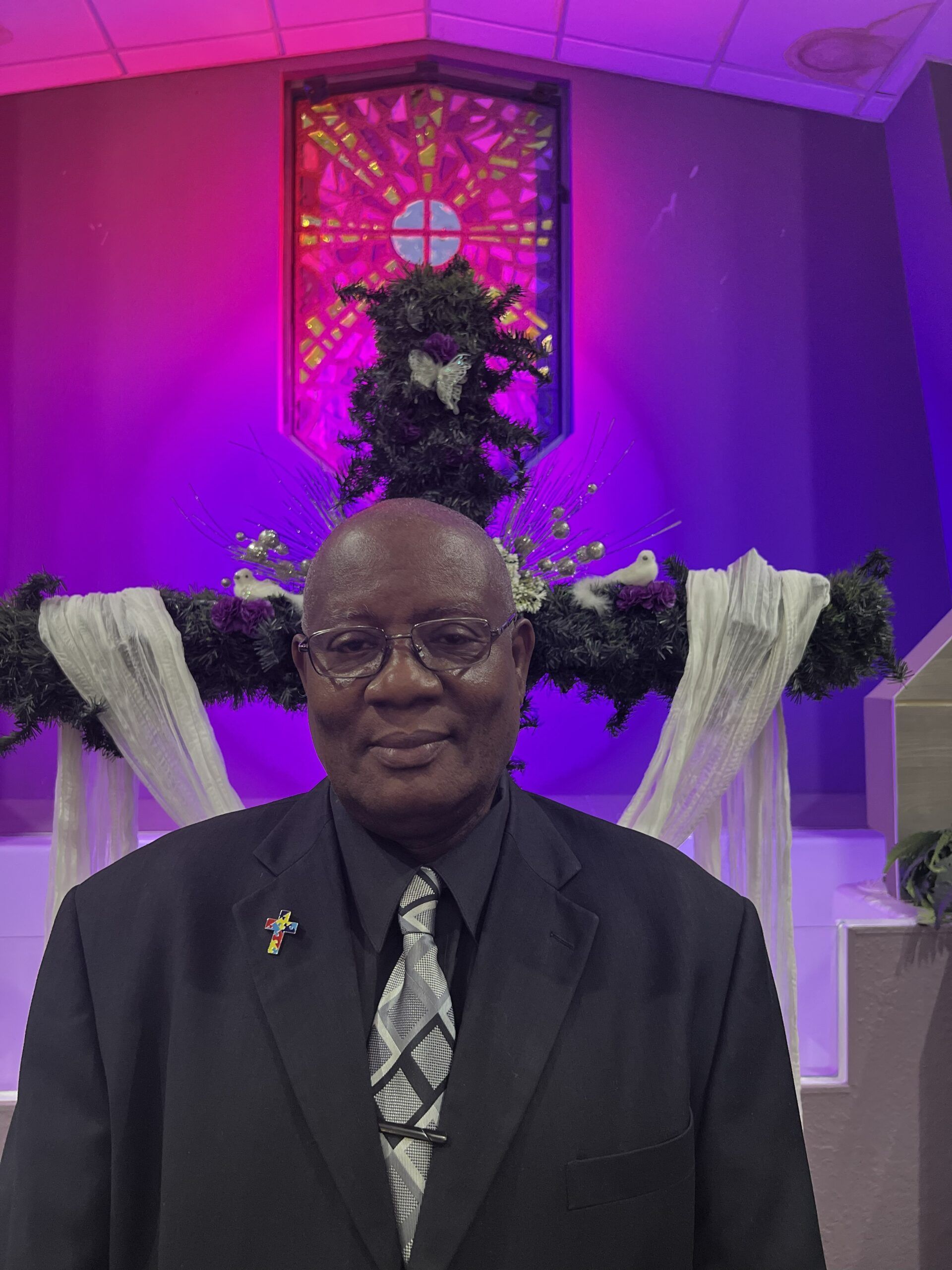 A person in a suit and patterned tie stands in front of a church altar decorated with dark flowers and purple lighting.