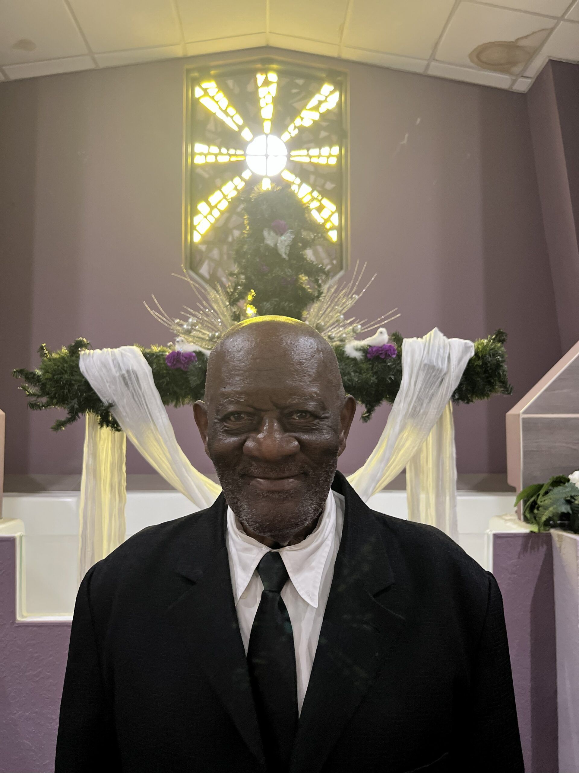 A person in a black suit and white shirt standing in front of a church altar decorated with flowers and a cross.