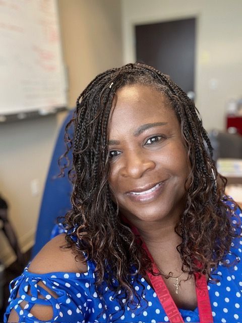 A smiling person with braided hair wears a blue and white polka-dot off-the-shoulder top in an office setting.