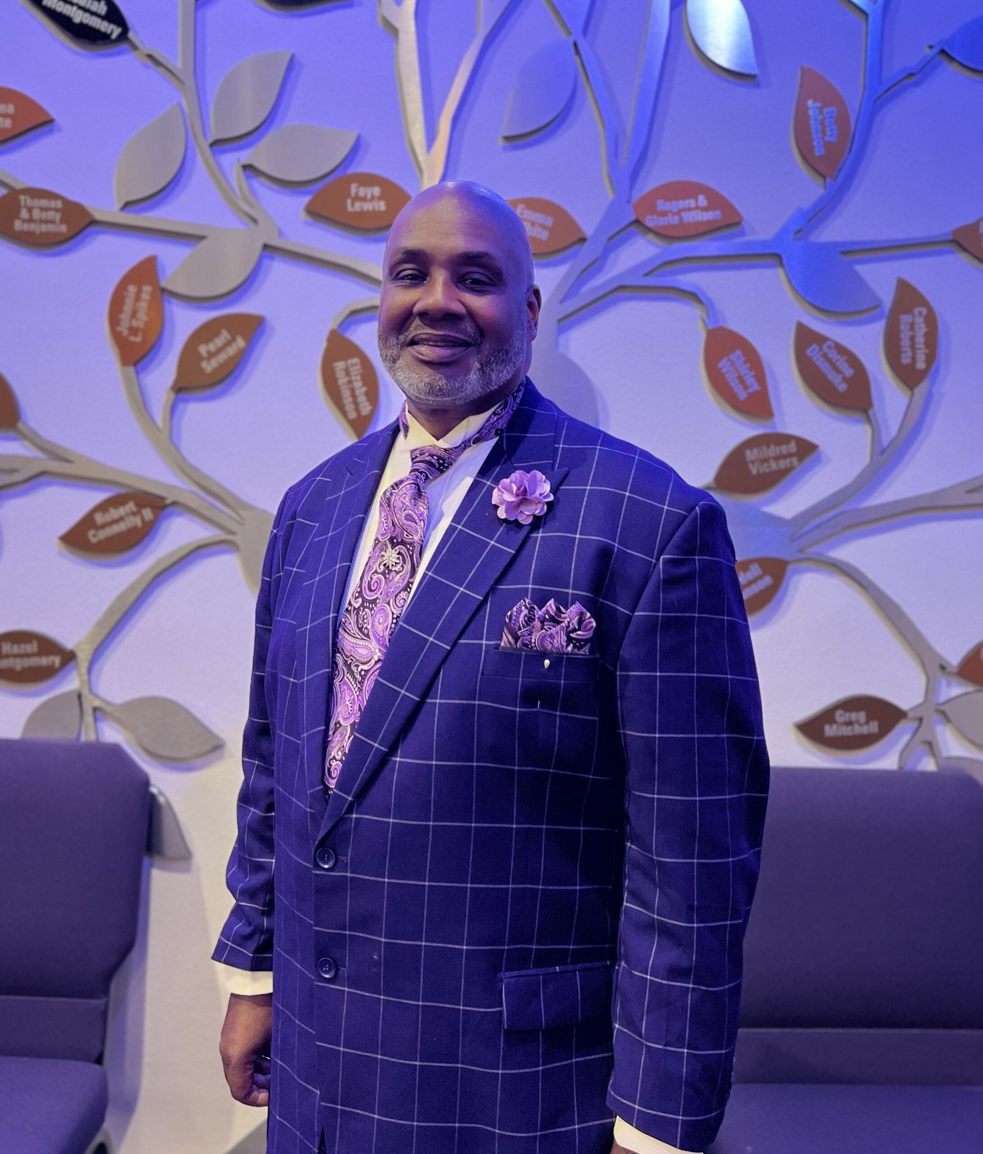 A person in a purple plaid suit and patterned tie stands in front of a wall decorated with a stylized tree and leaves.