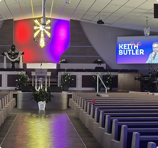 Interior of a church with rows of purple pews, a stage with floral arrangements, red and purple lighting, and a screen.