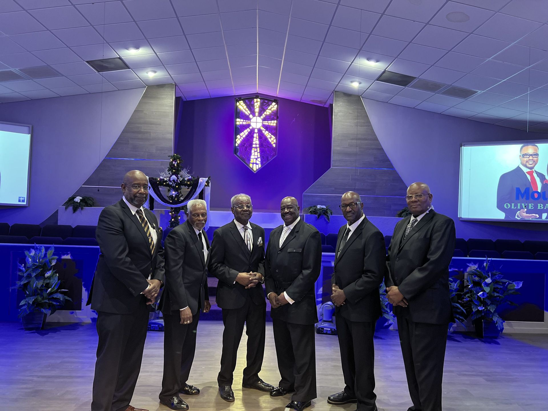 Six people in matching dark suits stand in a row on a stage in front of a purple-lit sanctuary backdrop.