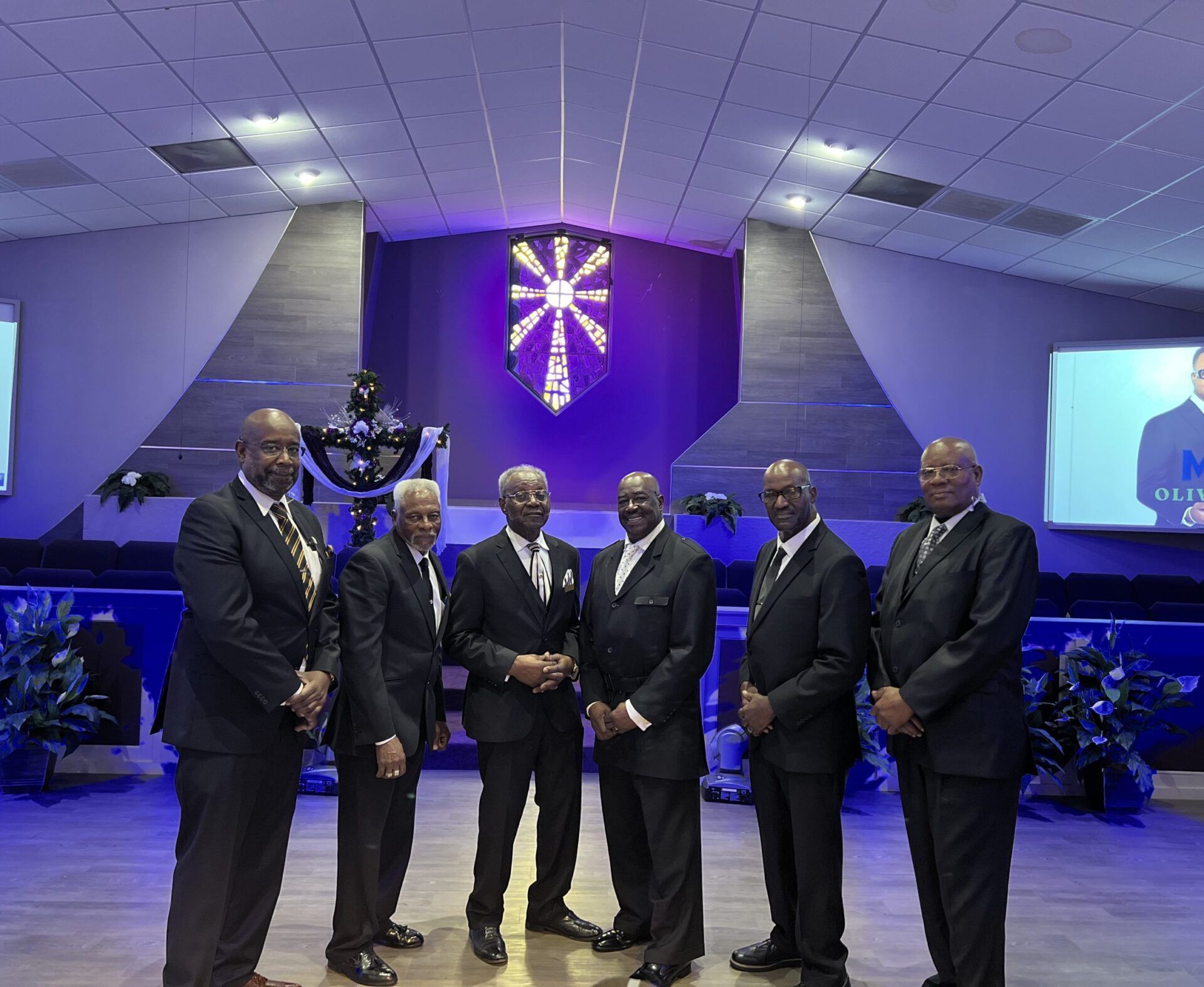 Six men in dark suits stand in a row on a church stage in front of a purple-lit cross, posing for a group portrait.