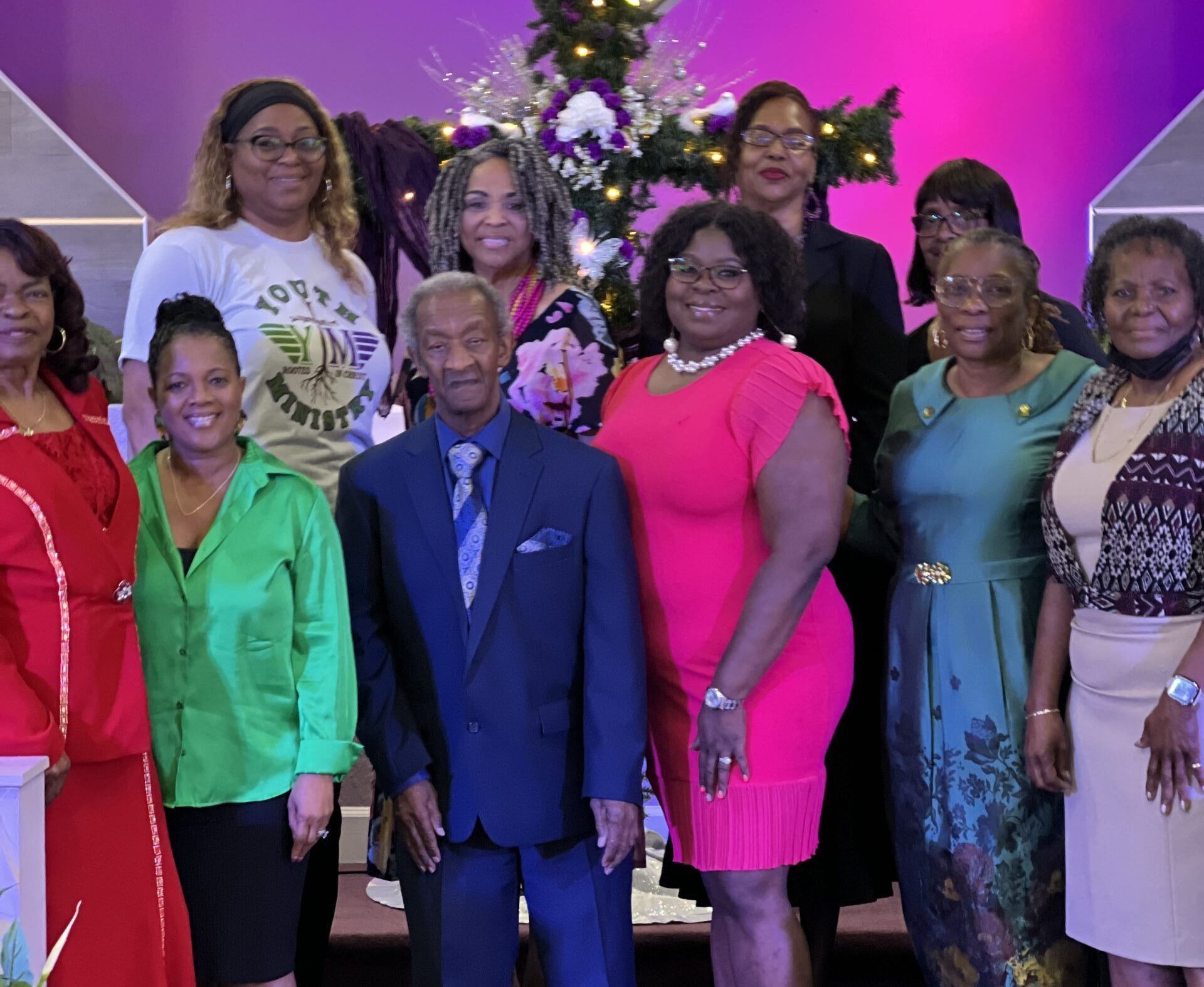 A group of ten people stand smiling for a photo in front of a decorated cross, wearing various formal and casual clothing.