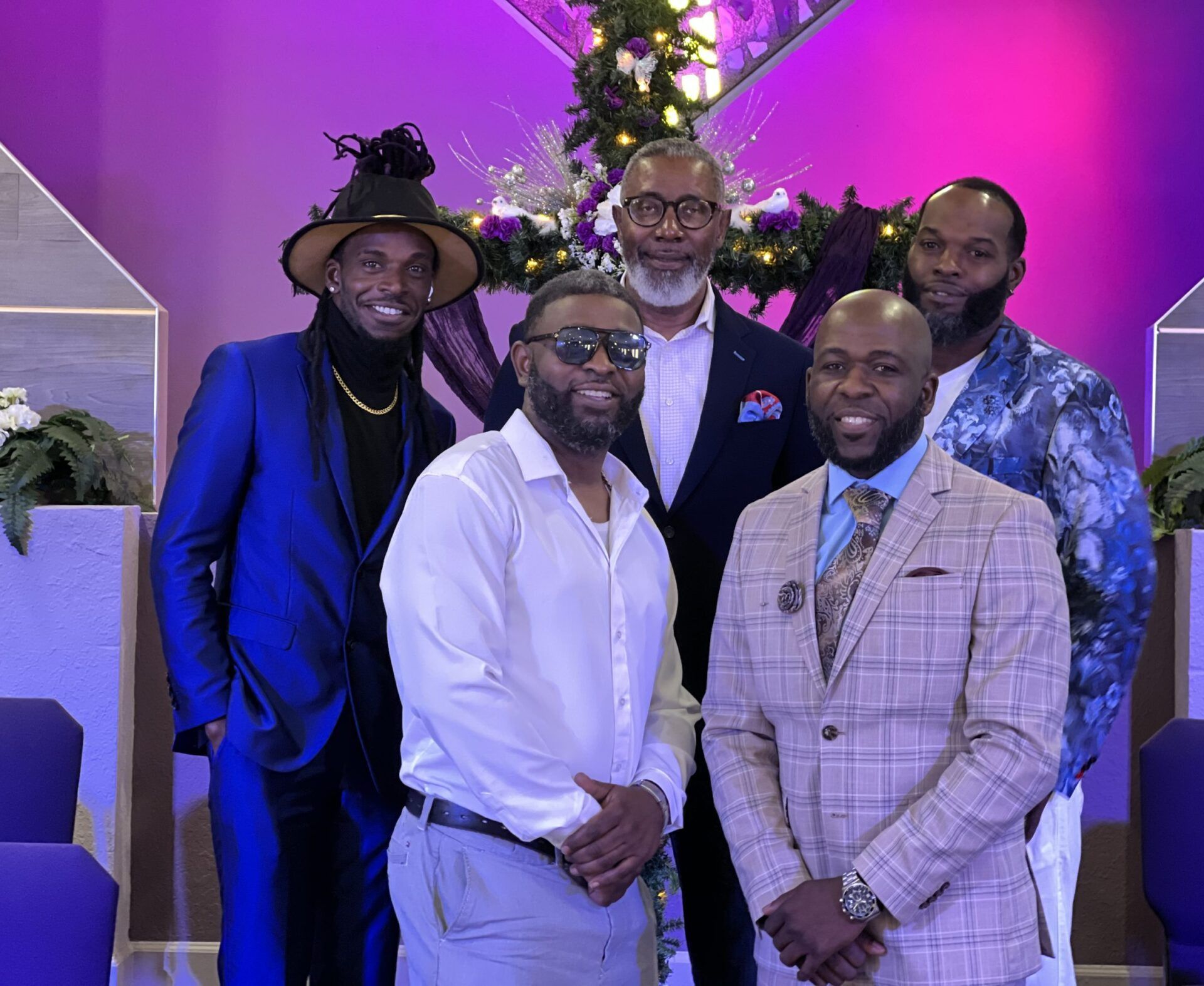 Five people stand together in a line, dressed in formal attire, inside a room with purple walls and holiday decor.