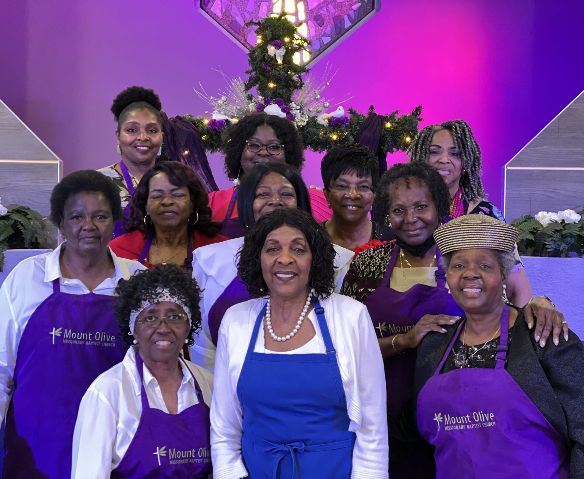 A group of people wearing purple aprons pose for a photo in front of a church altar decorated with greenery and lights.