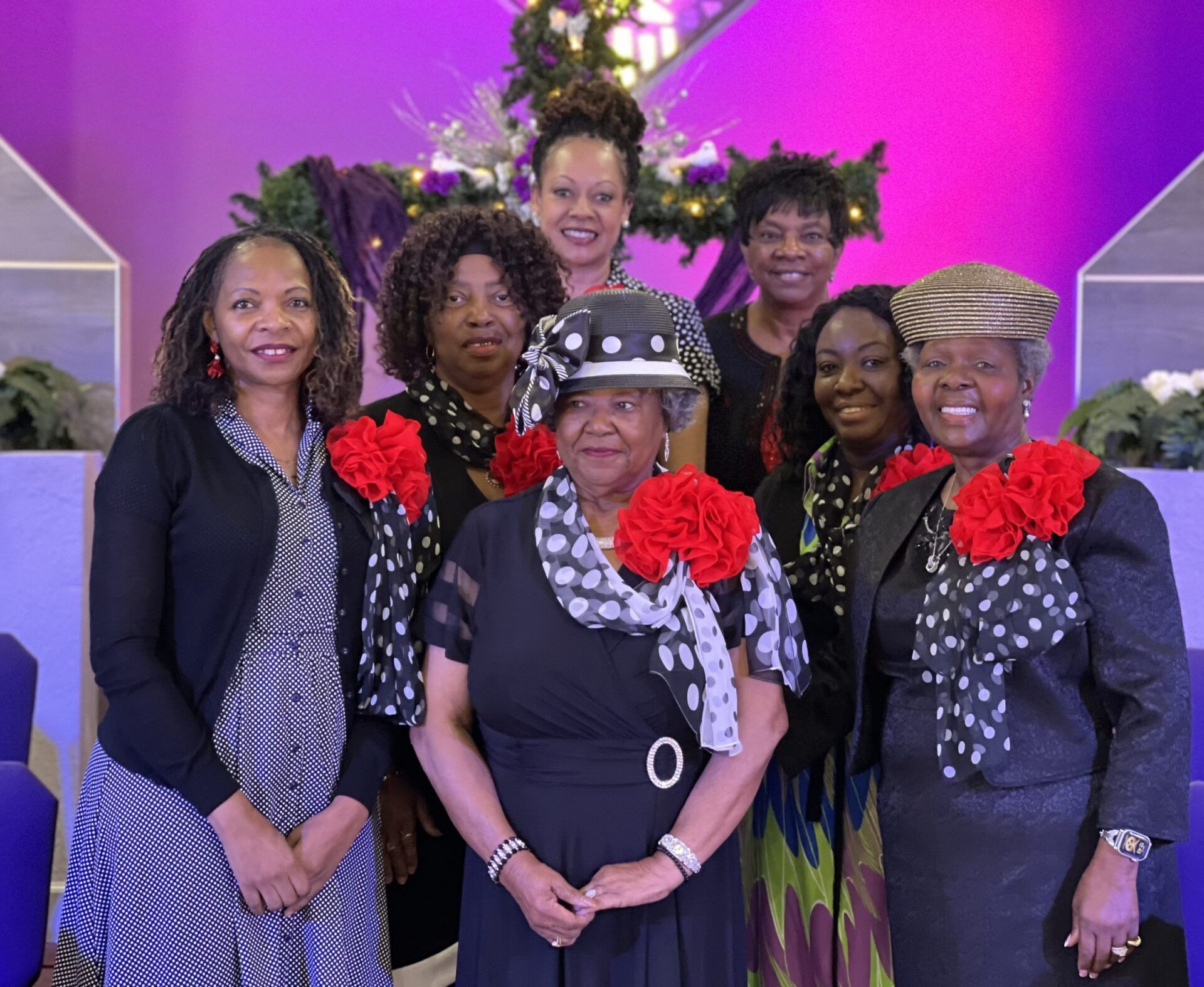 Seven people wearing coordinated black and white outfits with red accents pose together in a room with purple lighting.