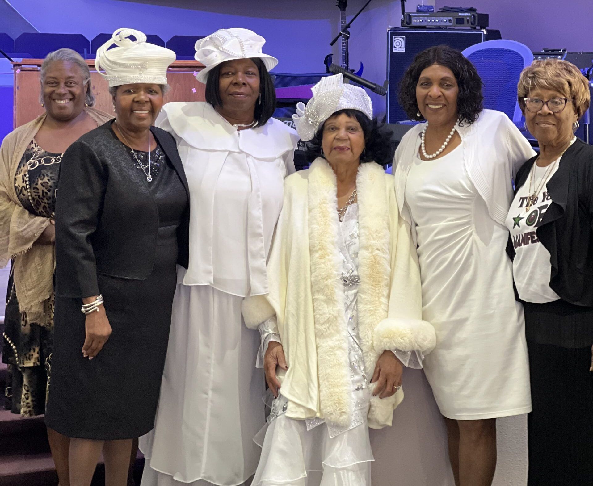 Six women in formal church attire stand together smiling in a room with musical equipment in the background.