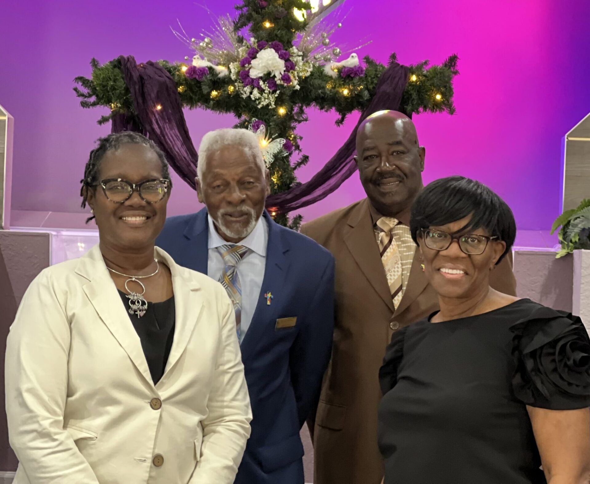 Four people pose smiling in front of a cross decorated with lights and flowers, set against a purple backdrop.