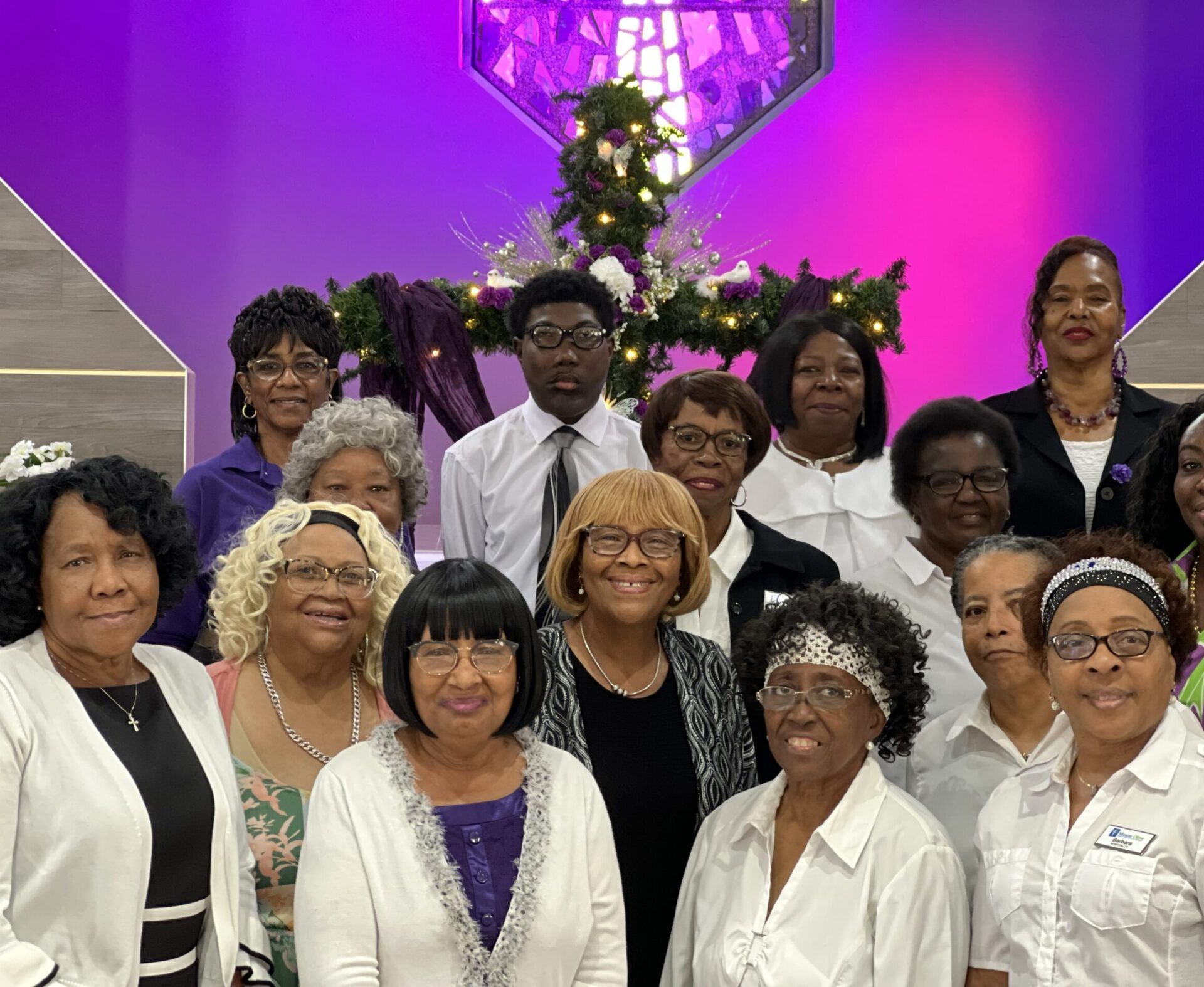 A group of smiling people stand together in front of a decorated cross, illuminated by bright purple accent lighting.
