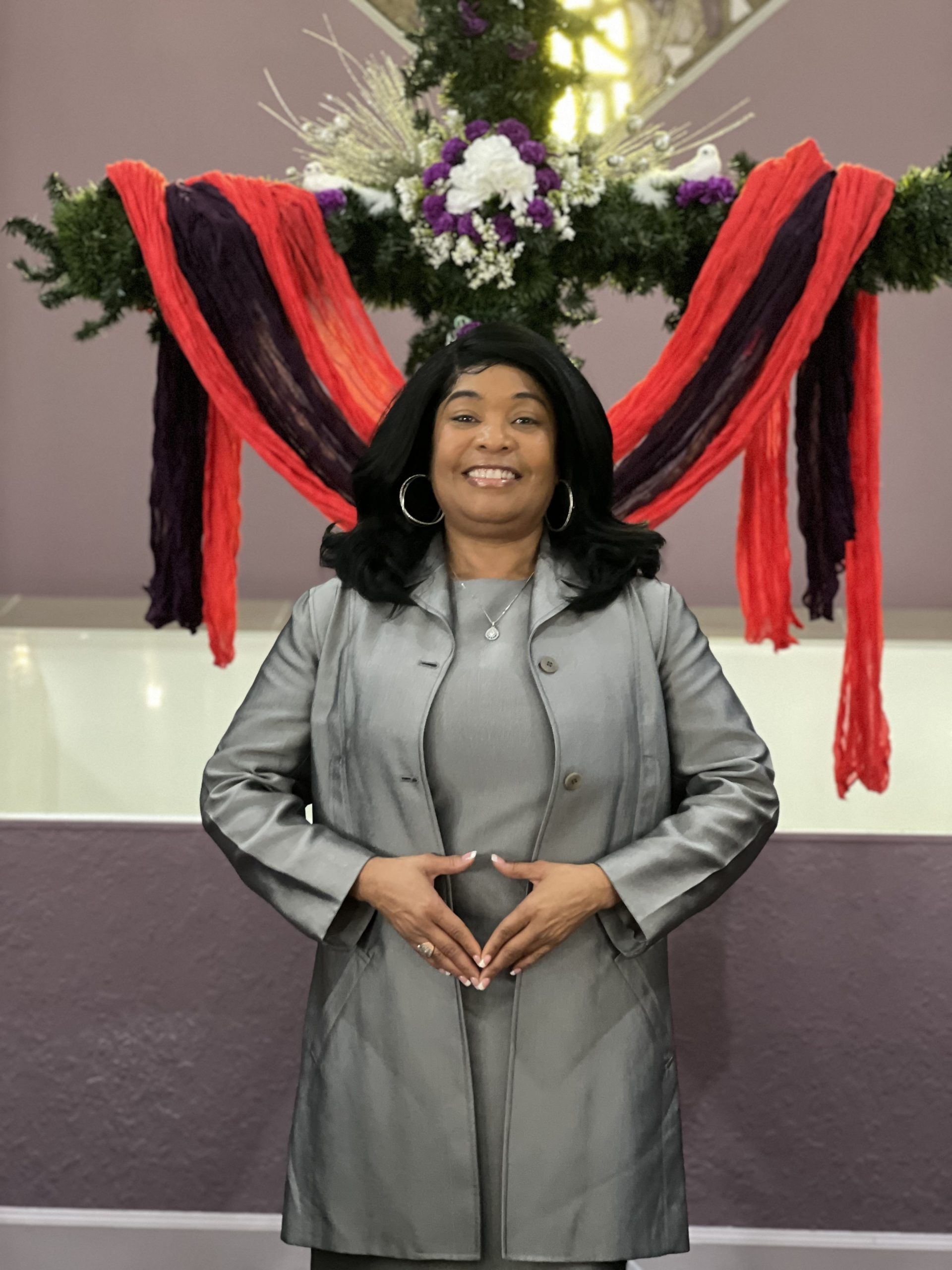 A smiling person wearing a gray suit standing in front of a cross adorned with red and purple fabric.