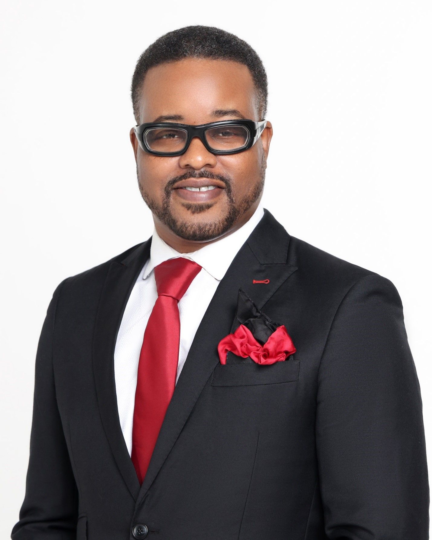 A person wearing glasses, a black suit, a white shirt, a red tie, and a red pocket square against a white background.