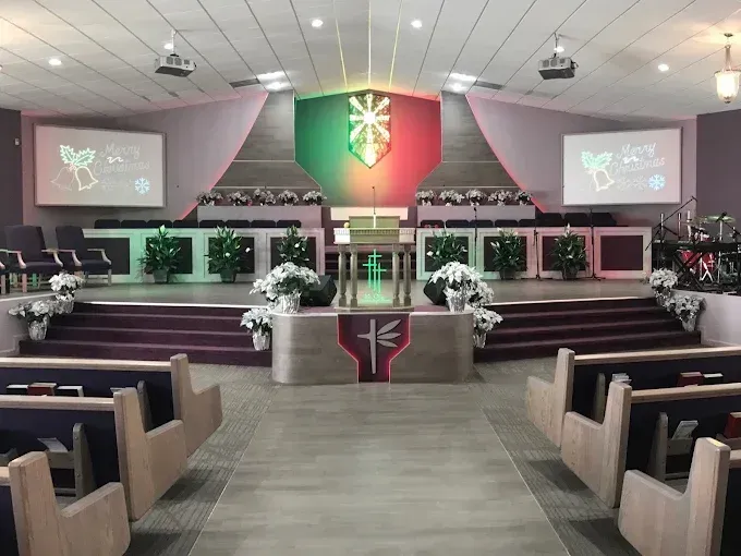 A modern church interior with rows of pews, a raised stage, floral arrangements, and two large projector screens.