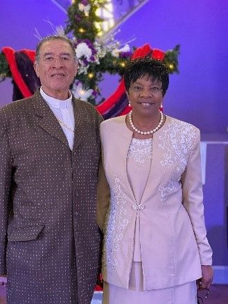 A couple stands together smiling, wearing formal attire, posed in front of a decorated cross against a purple background.