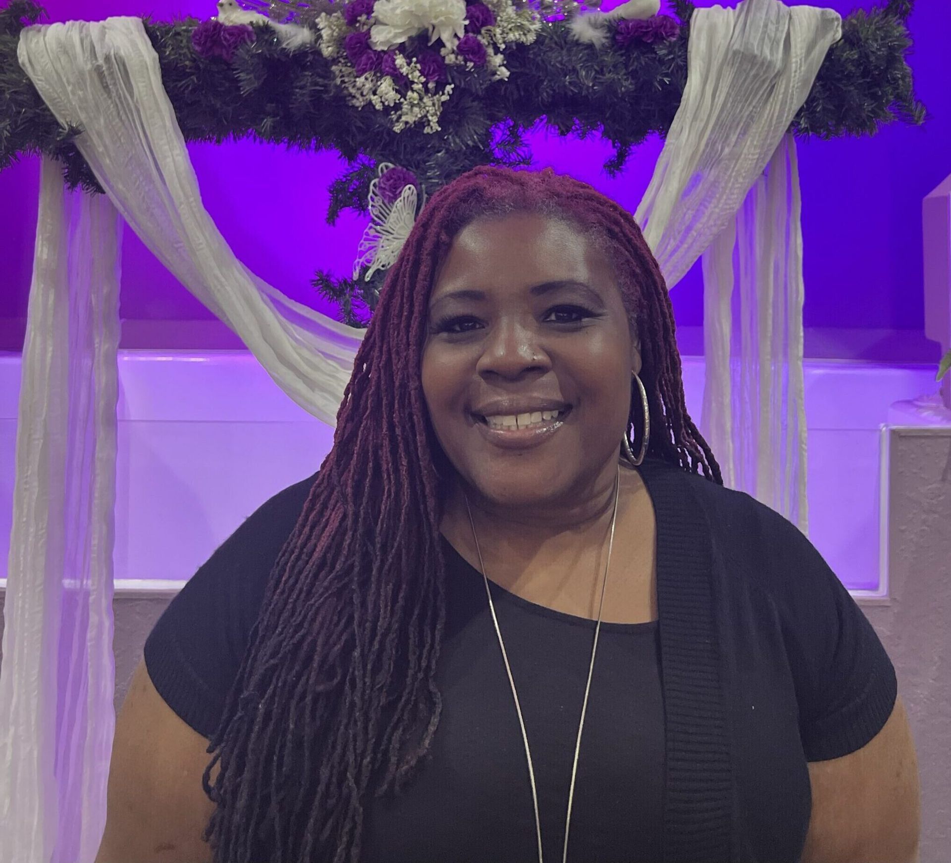 A smiling person with long auburn locs, wearing a black shirt, stands in front of a decorated cross with white fabric.
