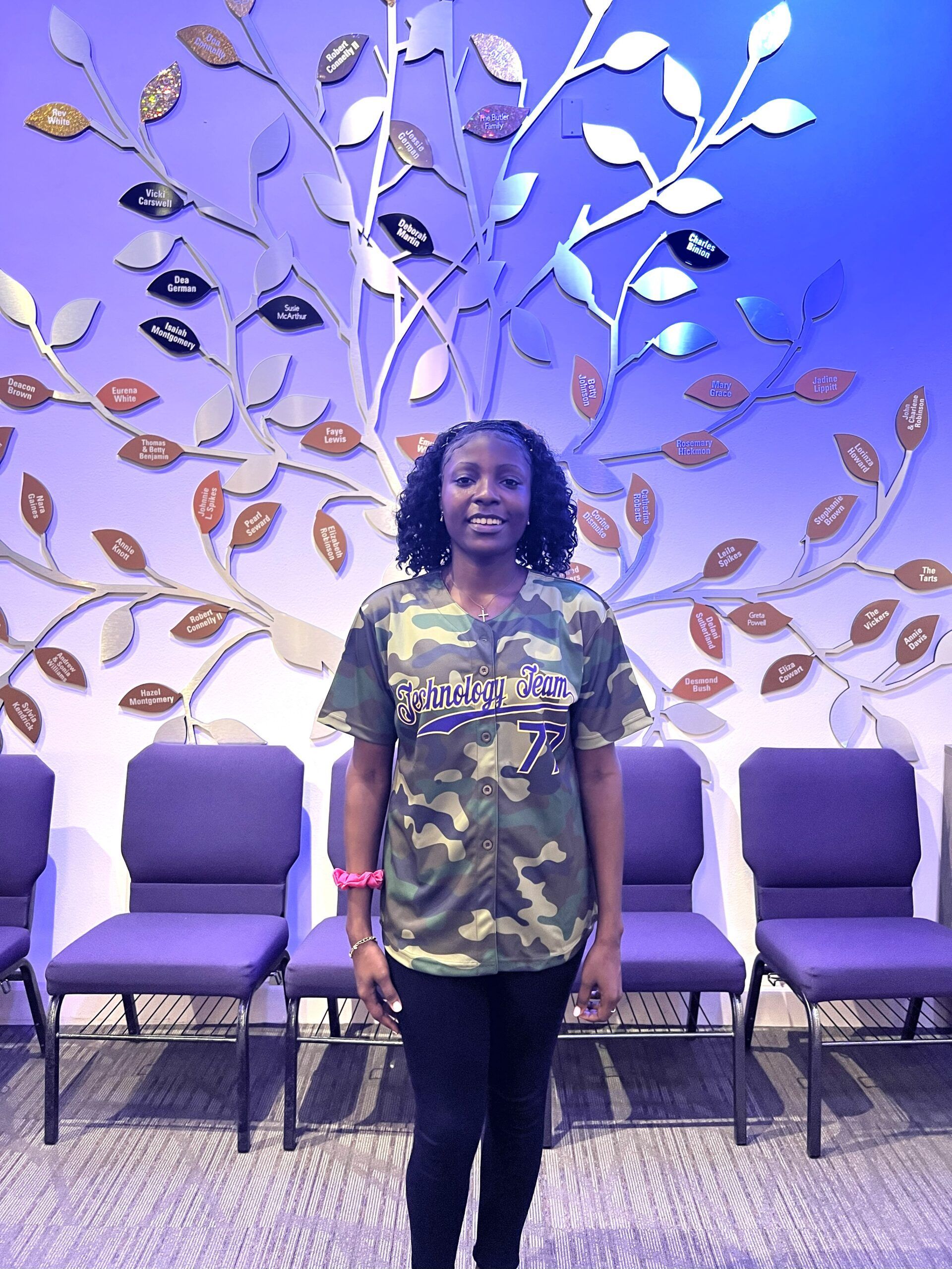 A person stands smiling in a camouflage shirt and black pants in front of a metal tree sculpture and purple chairs.