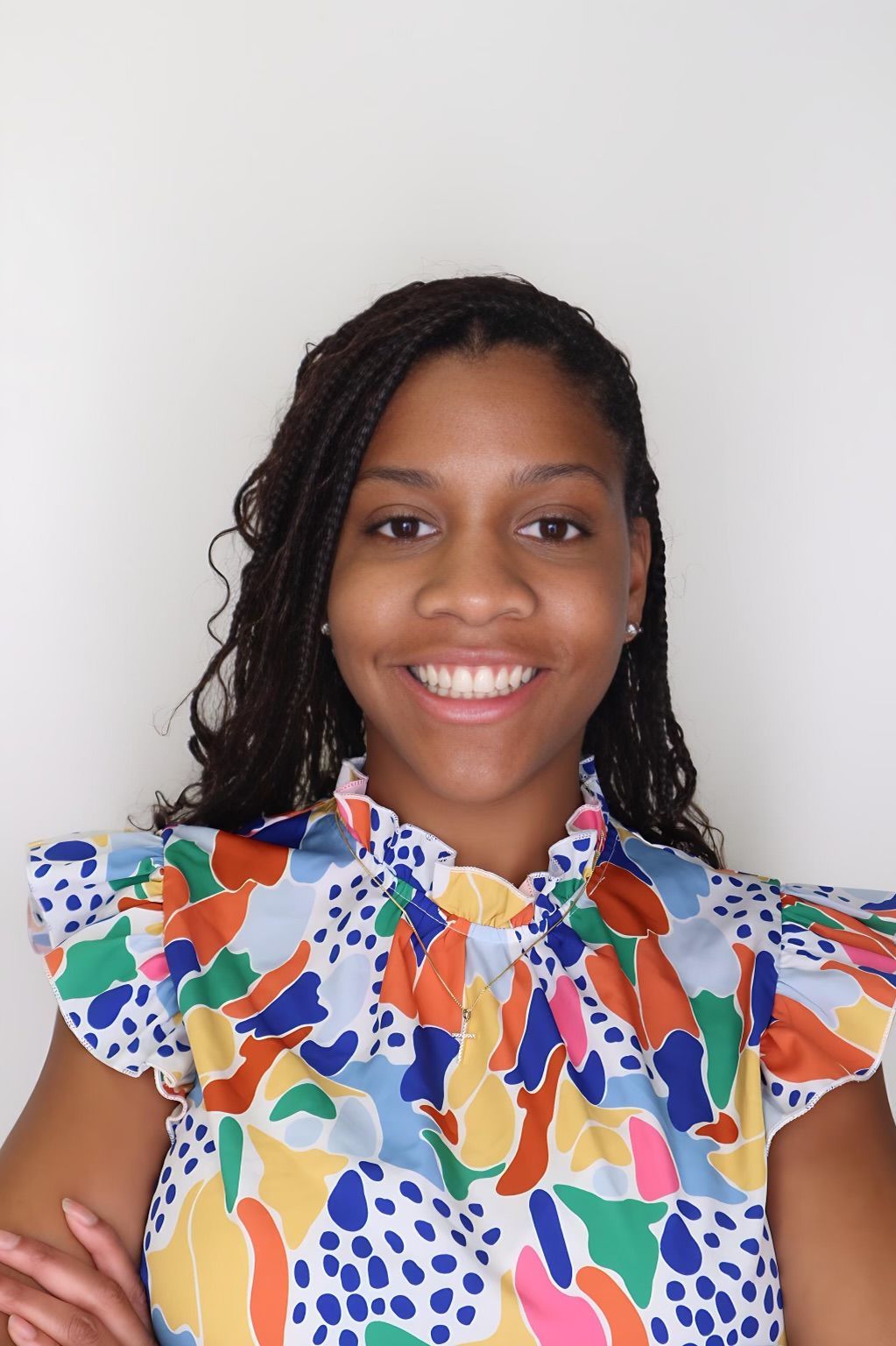 Smiling person with long, dark, braided hair wearing a sleeveless top with a colorful abstract print against a white wall.