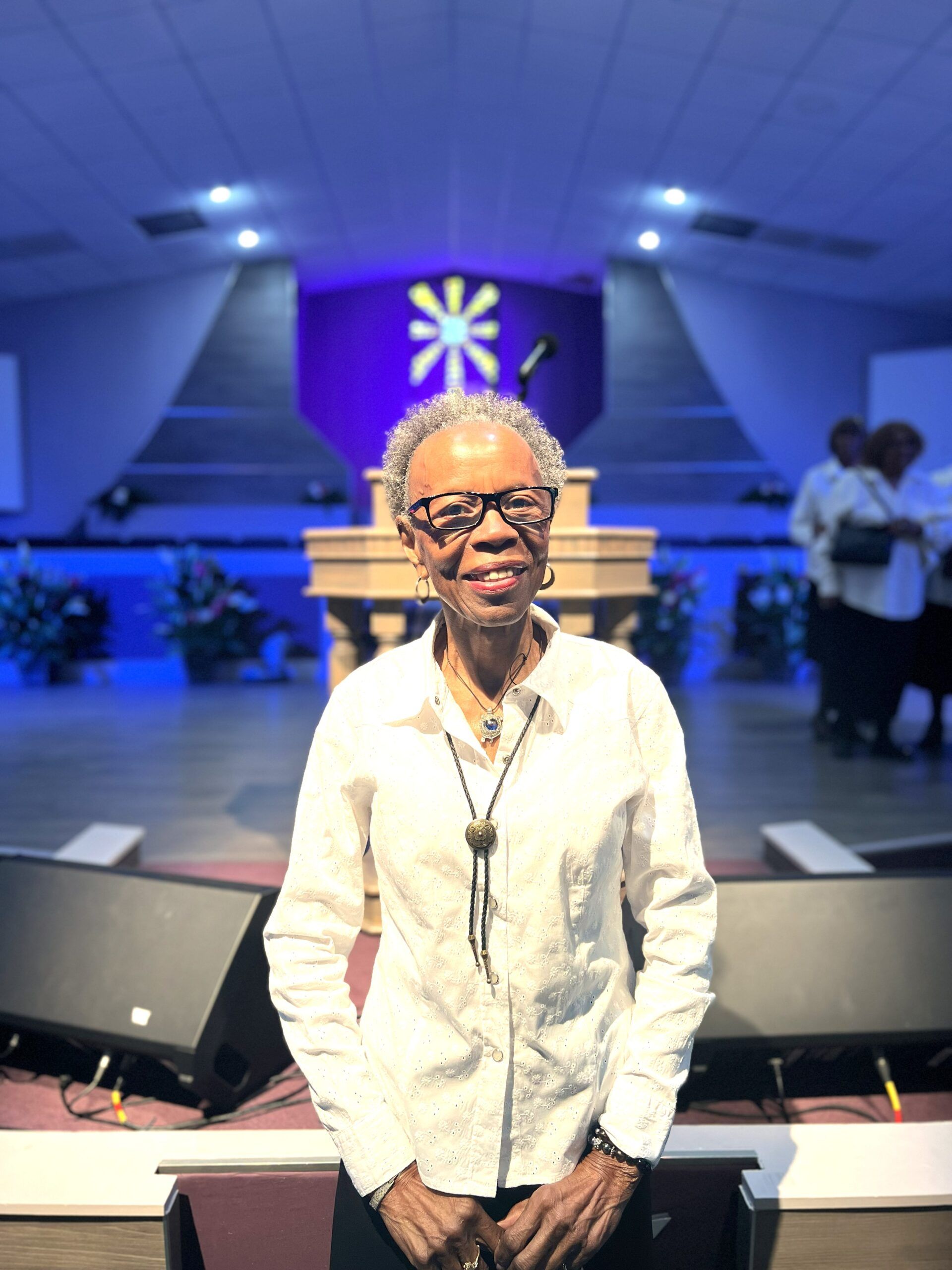 A person smiling while standing in front of a church pulpit with a purple illuminated background.