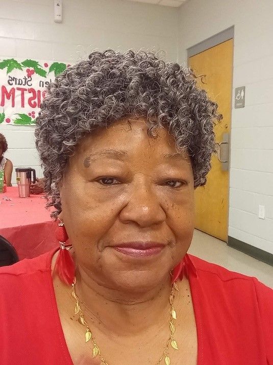 A person with short, curly, gray hair wears a red shirt, gold necklace, and red earrings in a room with a holiday sign.