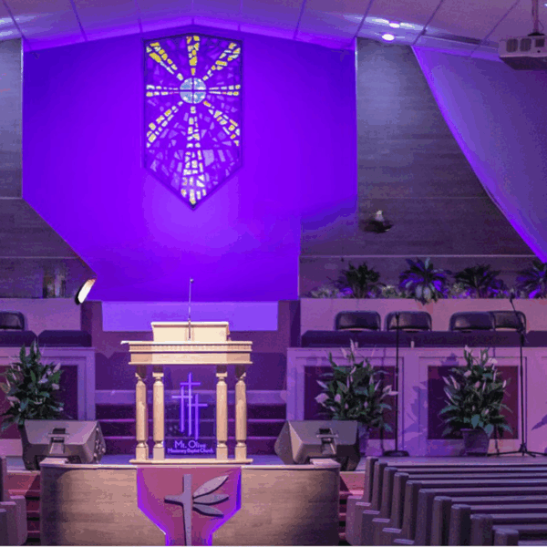 A wooden pulpit stands center stage in a church interior, illuminated by purple light, with a stained-glass cross above.