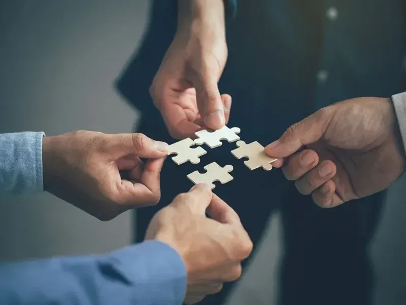 Four hands coming together to connect four small, light-colored puzzle pieces in a collaborative effort.