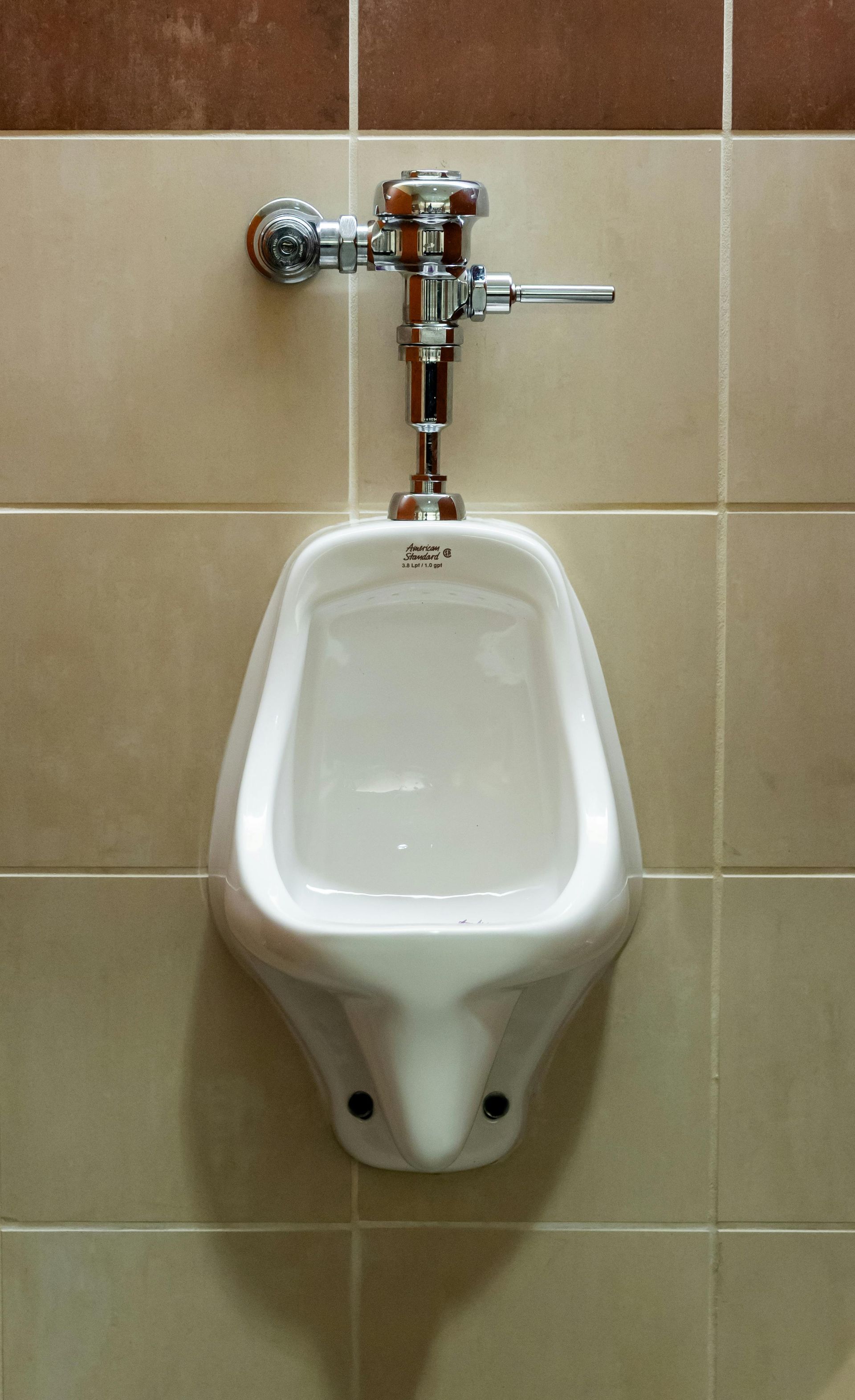 White urinal with chrome flushing mechanism mounted on beige tile wall.