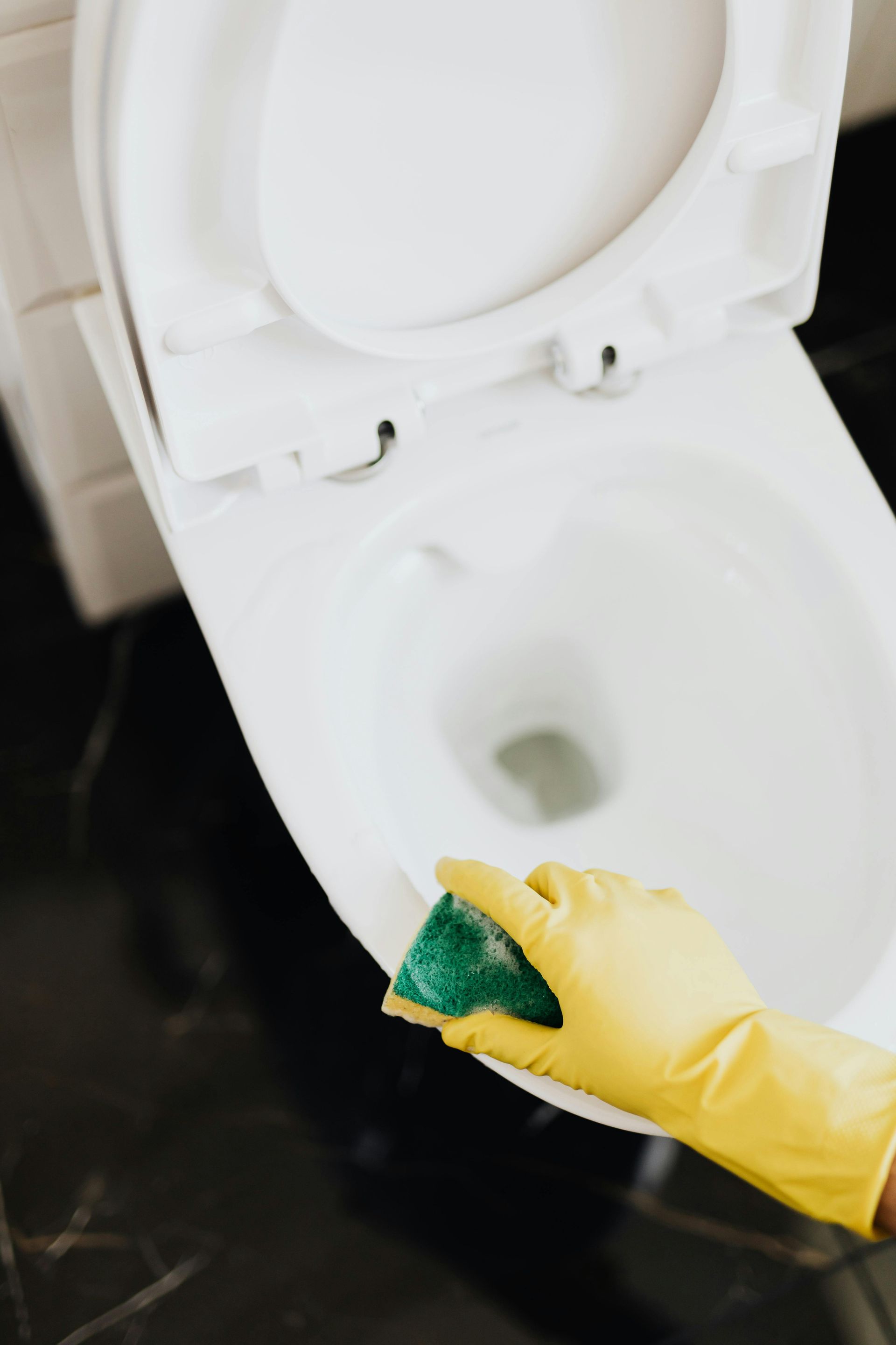 A person wearing a yellow glove cleans a white toilet with a green sponge after toilet install
