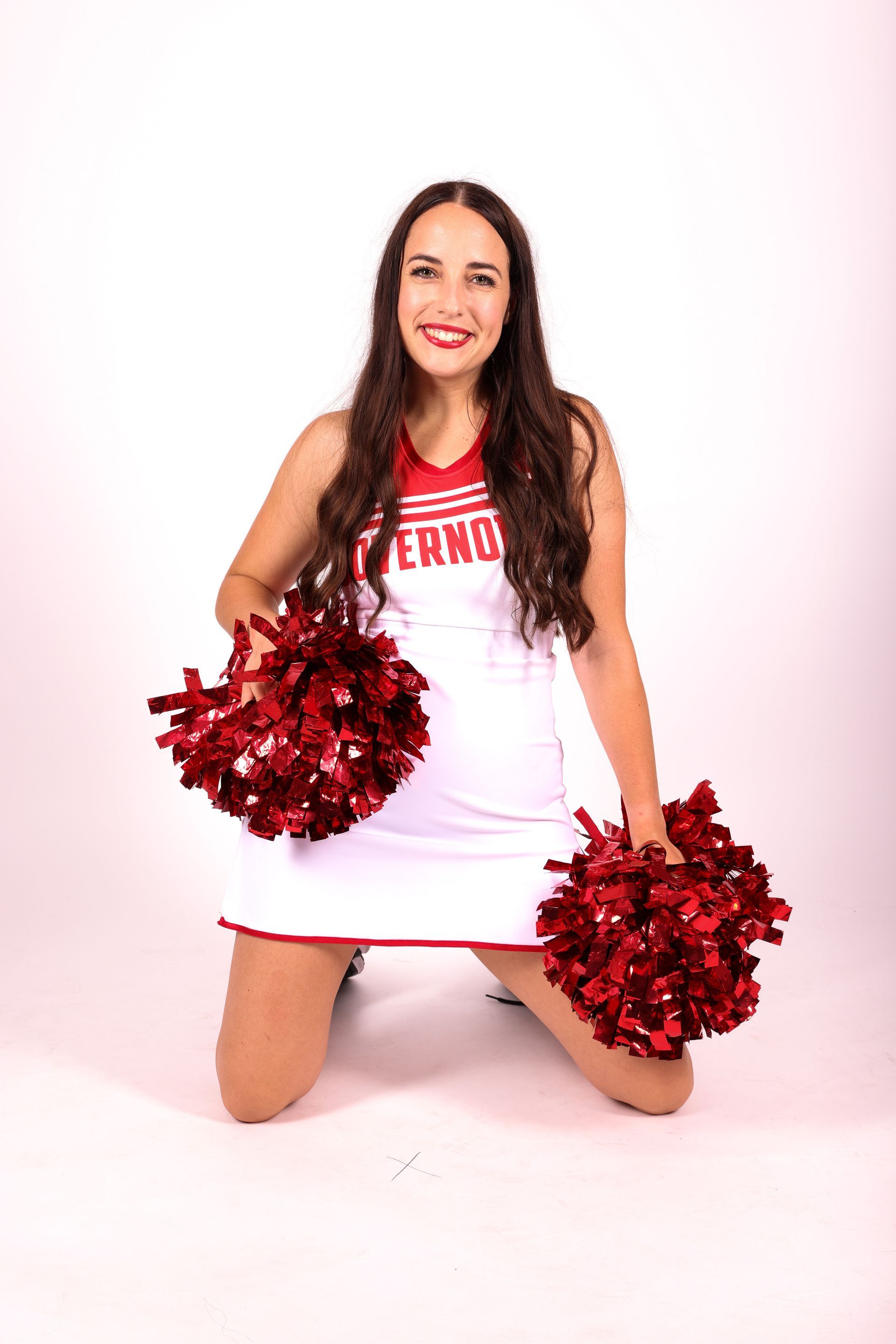 A cheerleader in a white dress is kneeling down holding red pom poms.