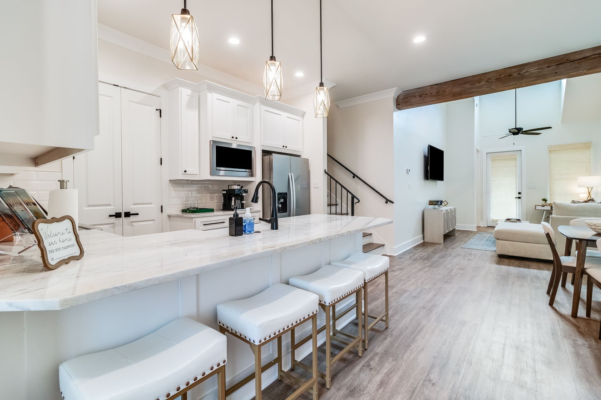 A kitchen with white cabinets , stools , a refrigerator and a staircase.