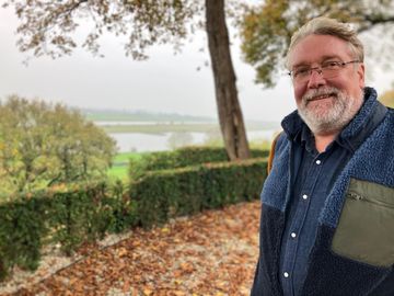 A man with a beard and glasses is standing in front of a lake.