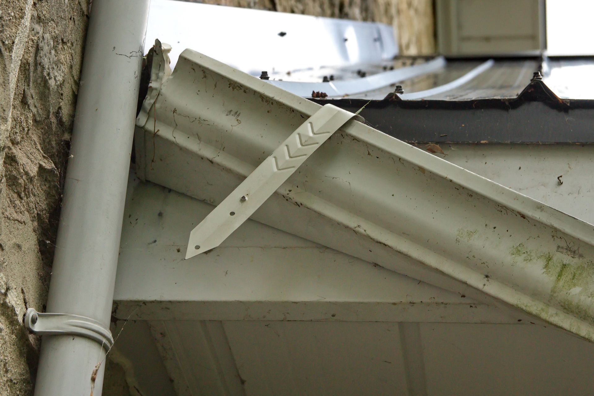 A detached white metal gutter bracket hangs loosely from the edge of a roof near a vertical downpipe and stone wall.