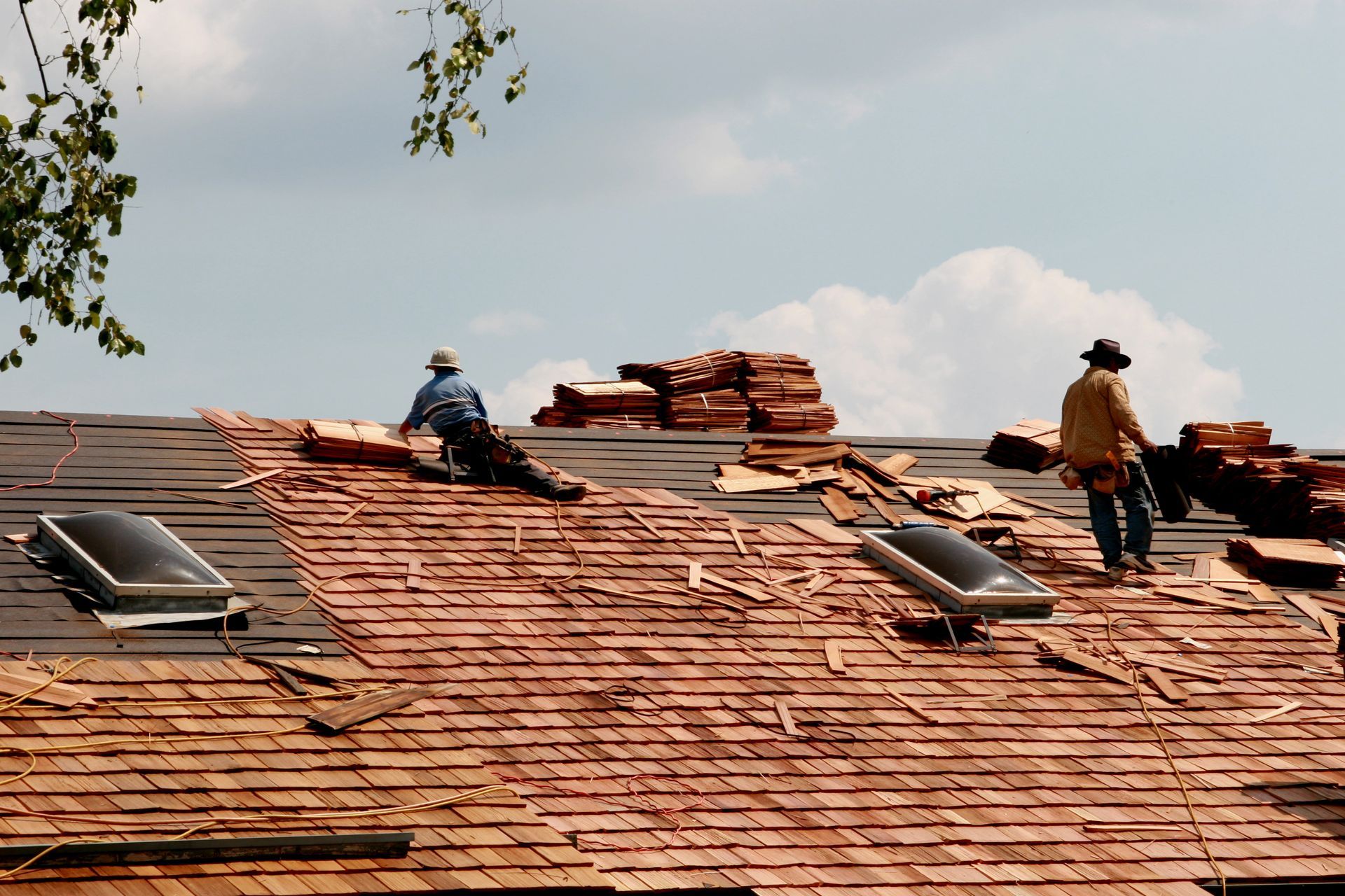 Two workers in hats replace clay tiles on a sloped roof with two skylights under a cloudy sky.