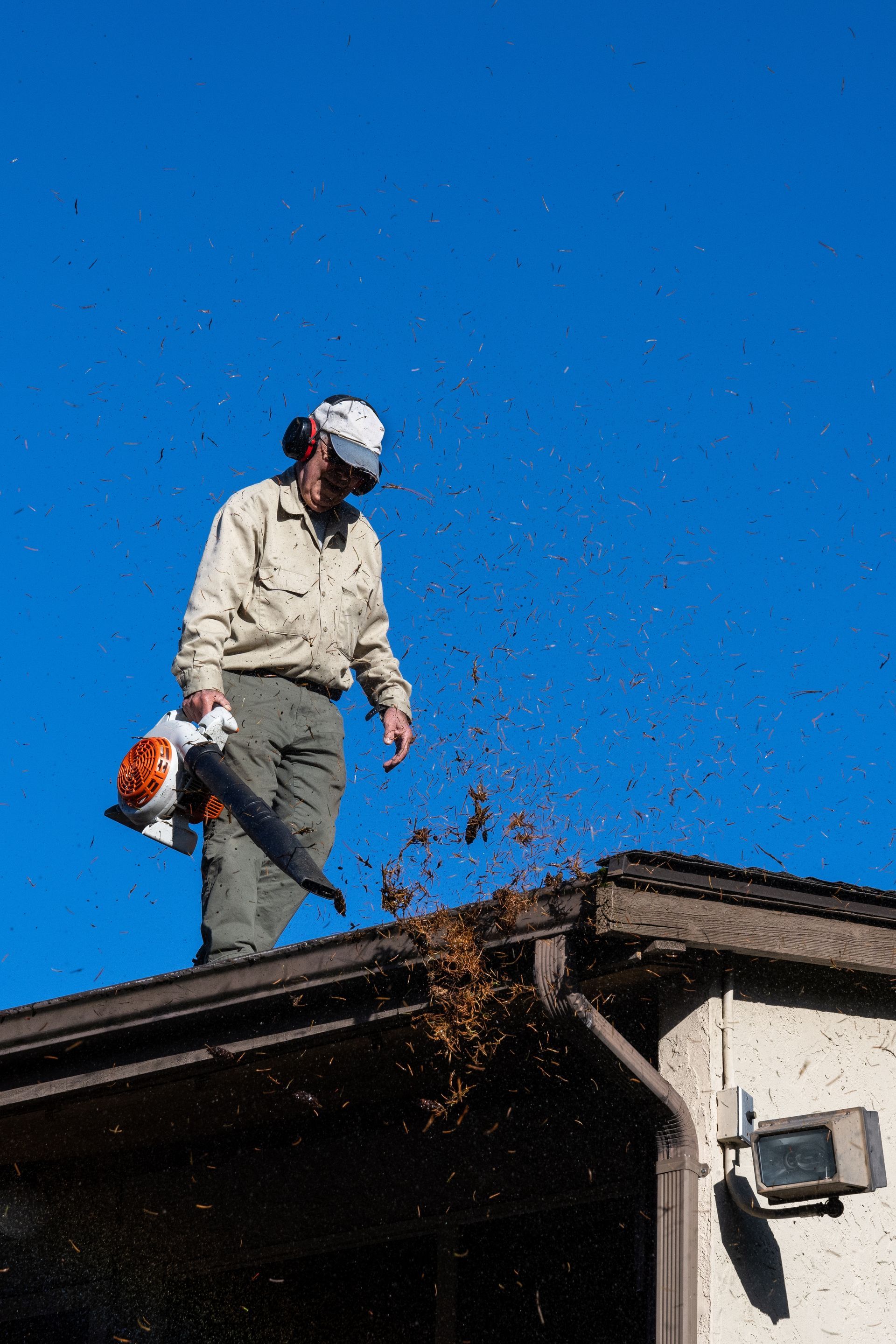 A person wearing ear protection and a cap uses a leaf blower to clear debris from a residential roof.